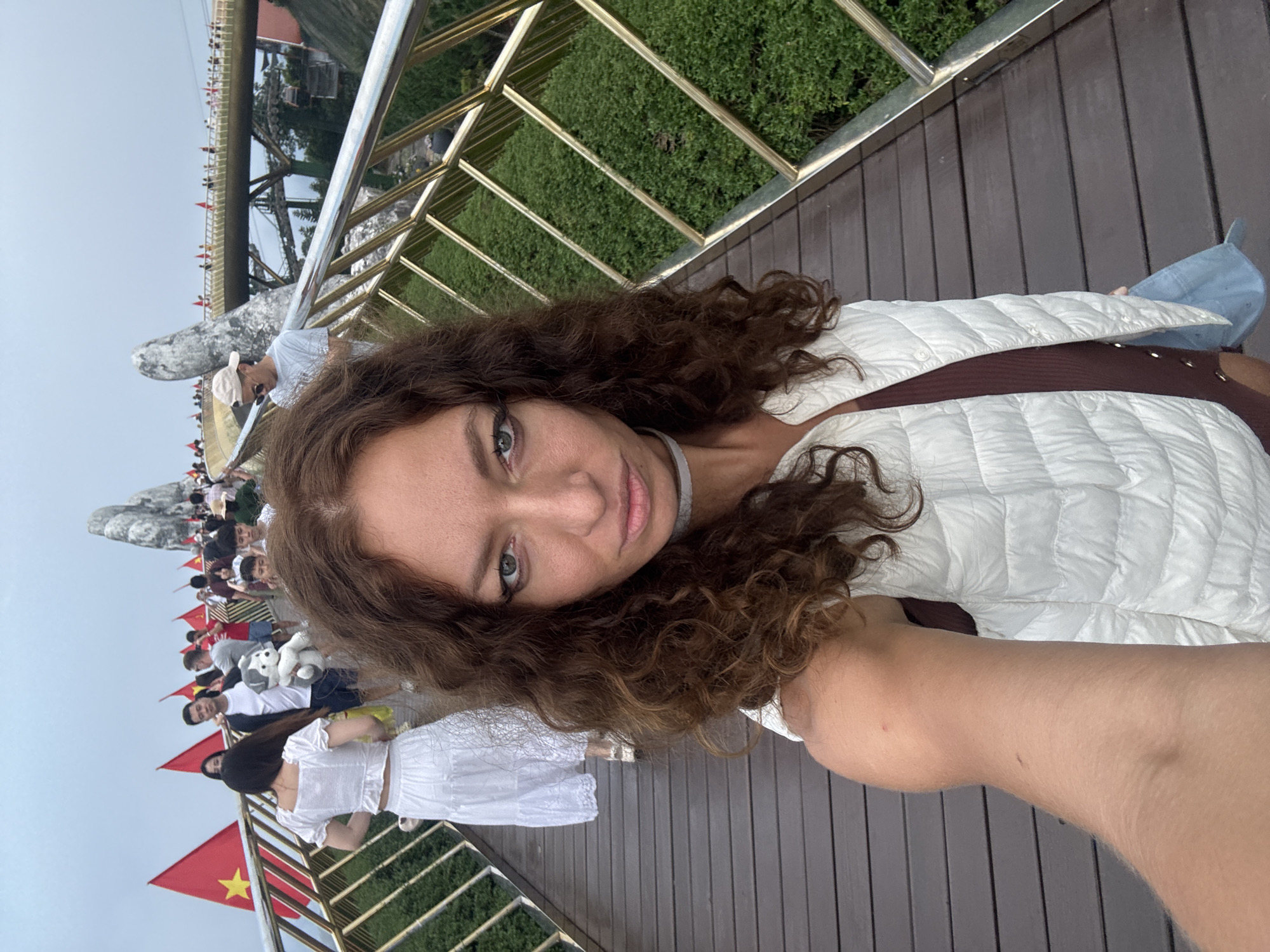 A selfie captures a woman with long, curly brown hair and light-colored eyes, looking directly at the camera with a neutral expression. She is wearing a white quilted jacket over a dark top and has a light-colored choker around her neck. Her right arm is visible, extended to take the photograph.
She is positioned on a wooden plank pathway, adjacent to a golden metal railing. In the background, numerous other people are visible walking along the pathway and on an adjacent structure. To the left, a distinctive large, grey, stone-like structure shaped like giant hands can be seen, which is a key feature of the Golden Bridge (Cầu Vàng) in Đà Nẵng, Vietnam. Further in the distance, what appears to be a metal truss bridge structure is visible under an overcast sky.
Many small red flags with a yellow star, representing the national flag of Vietnam, are placed along the bridge's railing. Below the railing to the right, green foliage is visible. The scene suggests a busy daytime tourist activity at a scenic location. No visible text is present in the image.