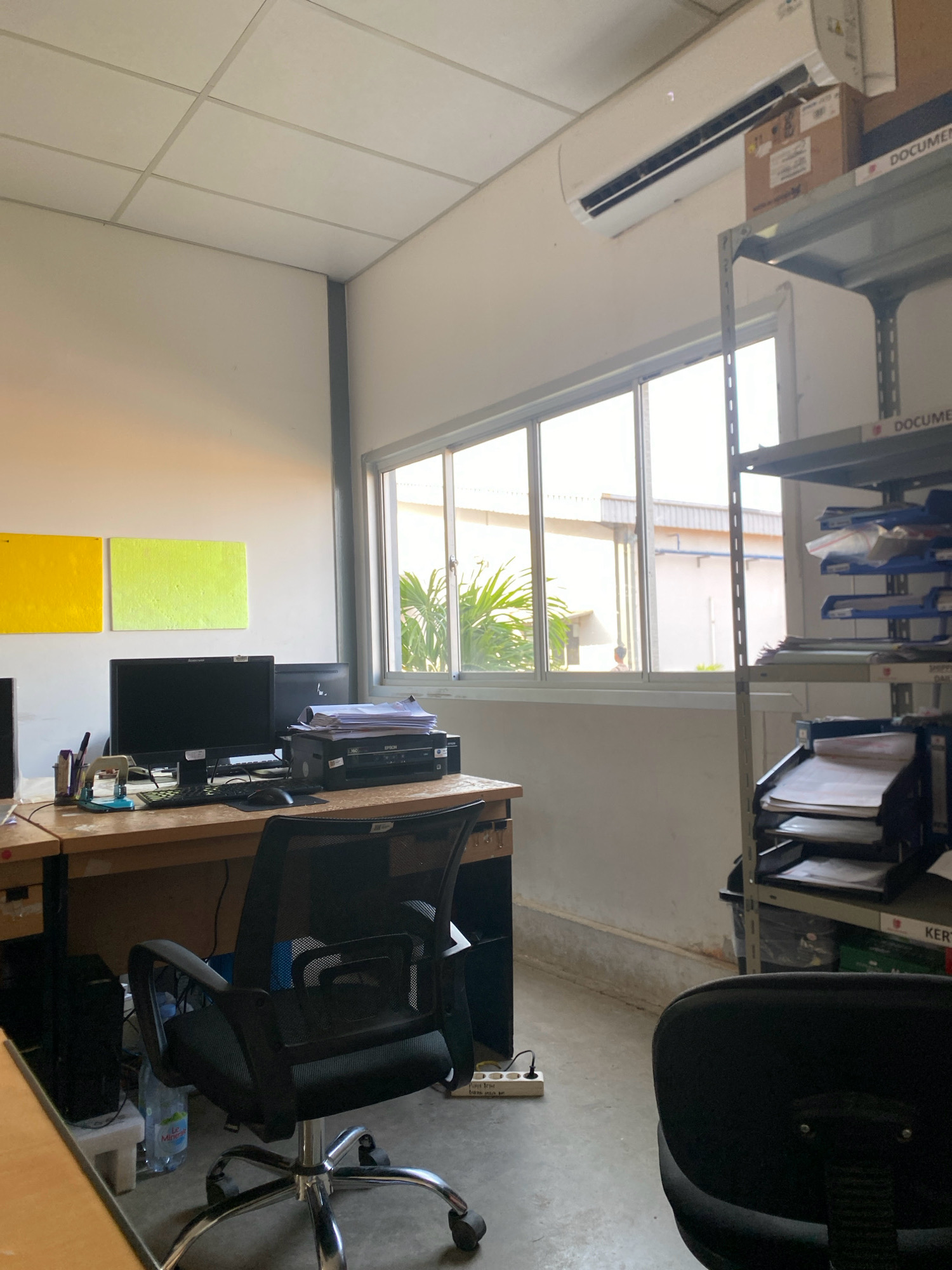 The image depicts a sparsely furnished office environment, possibly in Batam, Indonesia, characterized by its functional and utilitarian setup. The room features white walls and a suspended tiled ceiling with a visible air conditioning unit mounted high on one wall. A large window runs along one side, offering a view of outdoor greenery and a building with white walls, suggesting an industrial or commercial area.
The central focus of the image is a wooden desk equipped with two computer monitors, a keyboard, a mouse, and a printer. Office supplies such as pens, a stapler, and a hole punch are also present on the desk. An office chair with a black mesh back and seat is positioned in front of the desk. To the left of the desk, a black computer tower is visible, along with a bottle of water.
To the right of the desk, a tall metal shelving unit is filled with various office documents, folders, and storage boxes. Some of these items are labeled, with "DOCUMENT" and "KER..." partially visible, indicating organized storage. The floor is concrete, and a power strip with several outlets is visible near the desk.
Two rectangular canvases, one bright yellow and the other lime green, are mounted on the wall to the left of the desk, adding a touch of color to the otherwise neutral environment. The overall impression is that of a working space designed for efficiency and practicality, with minimal decorative elements.