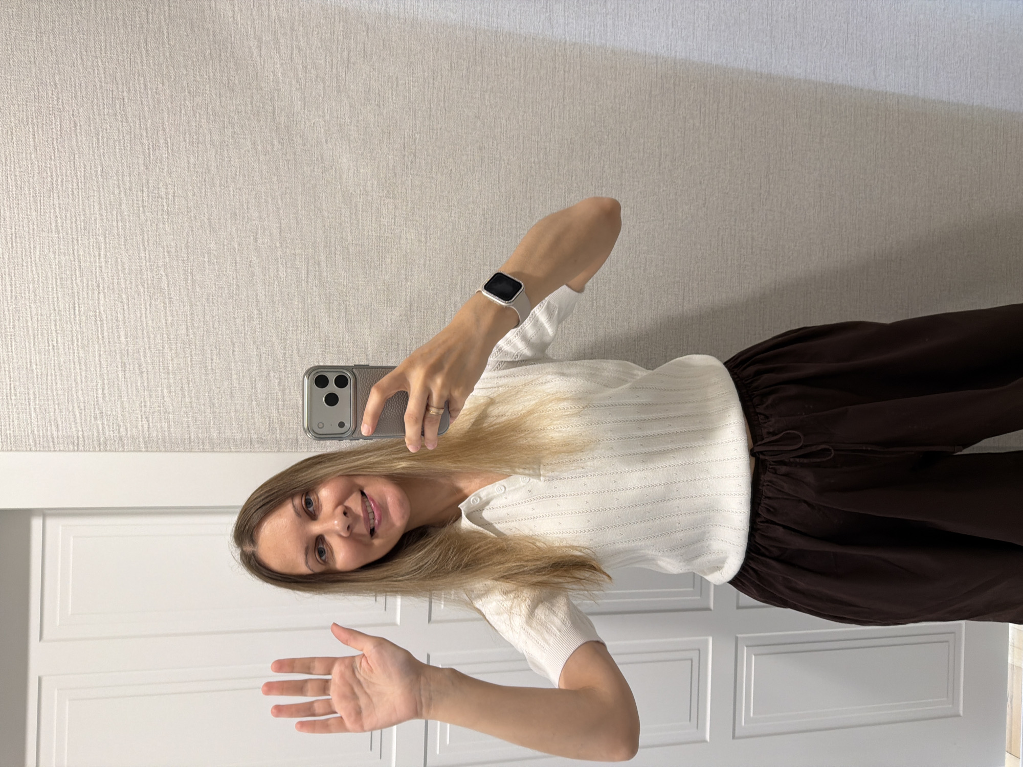 A woman is standing in what appears to be a hallway, possibly in a residential building, based on the double doors to her left and textured wallpaper to her right. She is holding a smartphone in her right hand, taking a selfie. Her left hand is raised, palm facing forward, in a waving gesture. She is smiling and looking at the phone's screen. The woman is wearing a white, short-sleeved collared shirt with a subtle textured pattern and dark brown wide-leg pants with a drawstring. A silver ring is visible on her left ring finger, and a smartwatch with a light-colored band is on her right wrist. The lighting suggests an indoor environment, possibly with natural light from an unseen source. The overall scene is casual and personal, captured in a mirror reflection or a selfie. There are no contextual clues to indicate this location is specifically in Izhevsk, Russia.