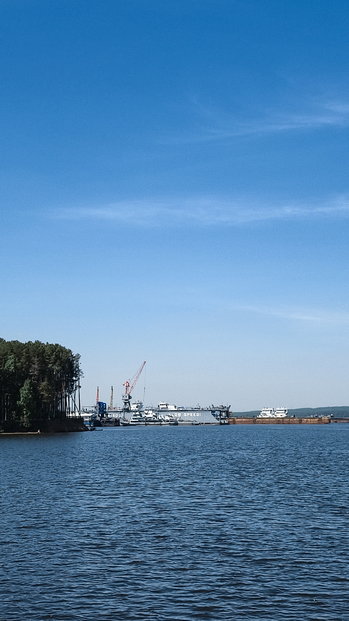 The image depicts a water view of a harbor or dock in Chaikovsky, Russia. A densely wooded shoreline is visible on the left edge of the frame. Several ships and barges are moored at the docks, with cranes and other industrial equipment visible. The phrase "SLOW SPEEDI" is visible on the side of a gray vessel. The water is dark blue and slightly choppy. A clear, blue sky with a wisp of clouds occupies the upper half of the image.