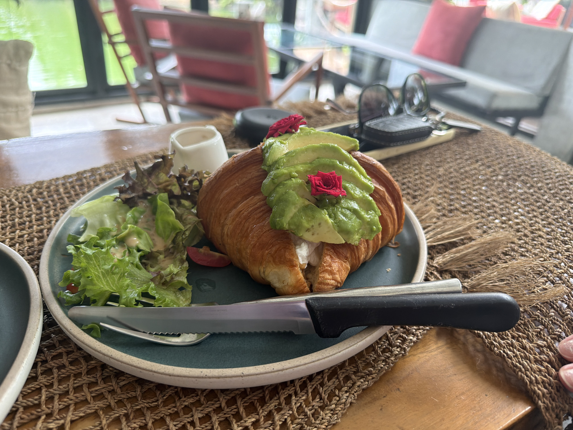 This media file shows a close-up of a croissant sandwich with avocado on a plate, part of a meal served in a restaurant in Baan Bo Phut, Thailand. The sandwich is topped with sliced avocado and two small red flowers. To the left of the sandwich is a small salad with lettuce and cherry tomatoes. A knife and fork are placed on the plate, ready for eating. In the background, a woven placemat and a wooden table are visible. Beyond the table, through a glass window, a lush green outdoor area with chairs and tables can be seen, suggesting an indoor-outdoor dining setting. The lighting suggests it is daytime. The overall ambiance appears relaxed and casual, typical of a cafe or restaurant setting.