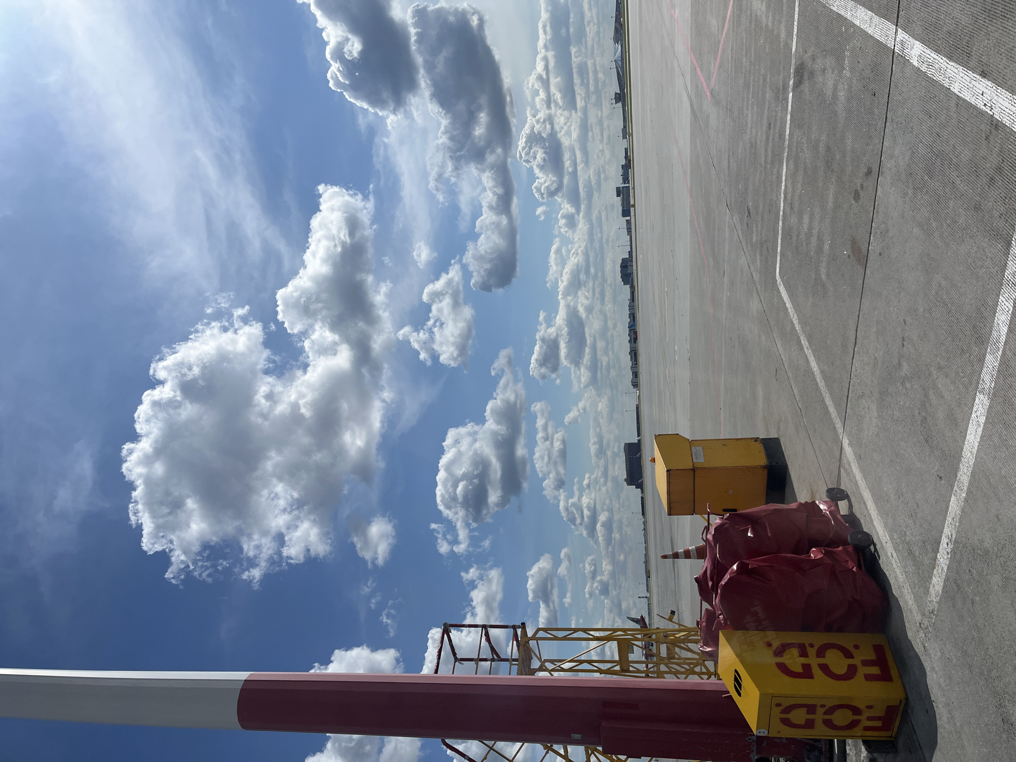 An outdoor medium shot captures an airport tarmac on a bright, partly cloudy day. In the foreground, a tall, red and white striped pole stands to the left, partially obscuring a yellow metal structure and two red-covered objects on wheels, both bearing yellow markings. A yellow bin with "FOD" stenciled in red is positioned to the left of the red-covered items. Further back, an orange traffic cone is visible, as well as another yellow bin. The background reveals a vast expanse of tarmac with red and white painted lines. In the distance, several airport buildings and structures are scattered across the flat landscape under a sky filled with white, puffy clouds. The scene is devoid of any people. The overall impression is of a typical operational area at an airport, possibly Sheremetyevo International Airport in Khimki, Russia, given the context.