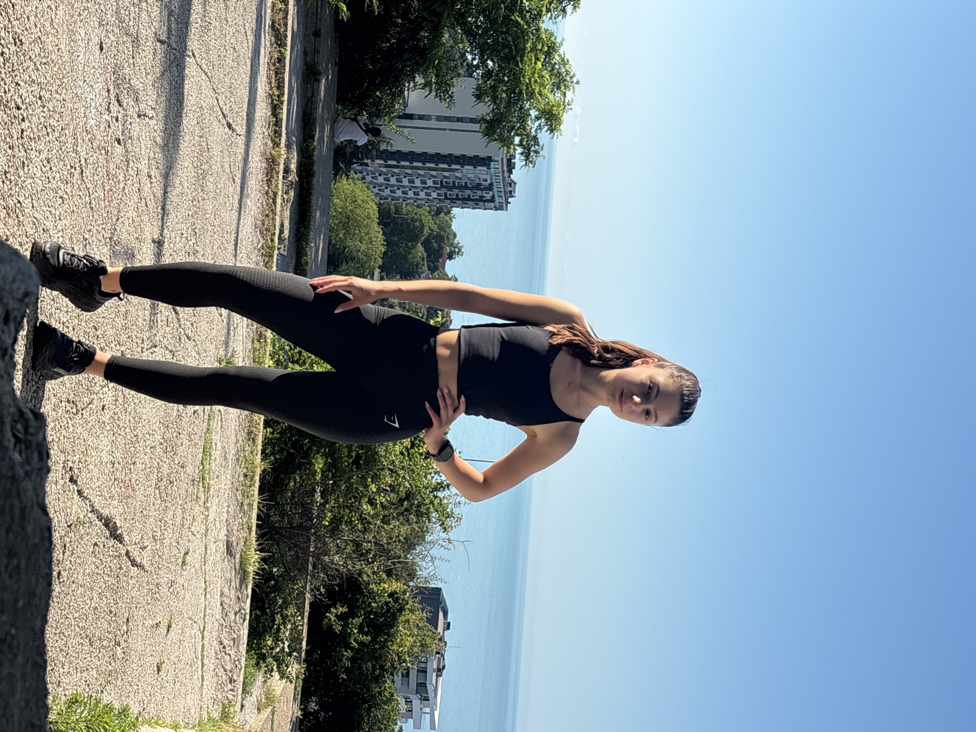 A young woman with dark hair pulled into a ponytail stands in athletic wear, a black crop top and black leggings, in what appears to be an outdoor setting. She is facing the camera with her left hand on her hip. In the background, there is a body of water, possibly the Black Sea, with buildings and lush greenery visible along the coastline. The ground in the foreground is paved and textured, with shadows cast from unseen objects. The sky is clear and blue. The location is likely a park or elevated viewpoint in the city of Varna, Bulgaria, offering a panoramic view of the sea and urban landscape.