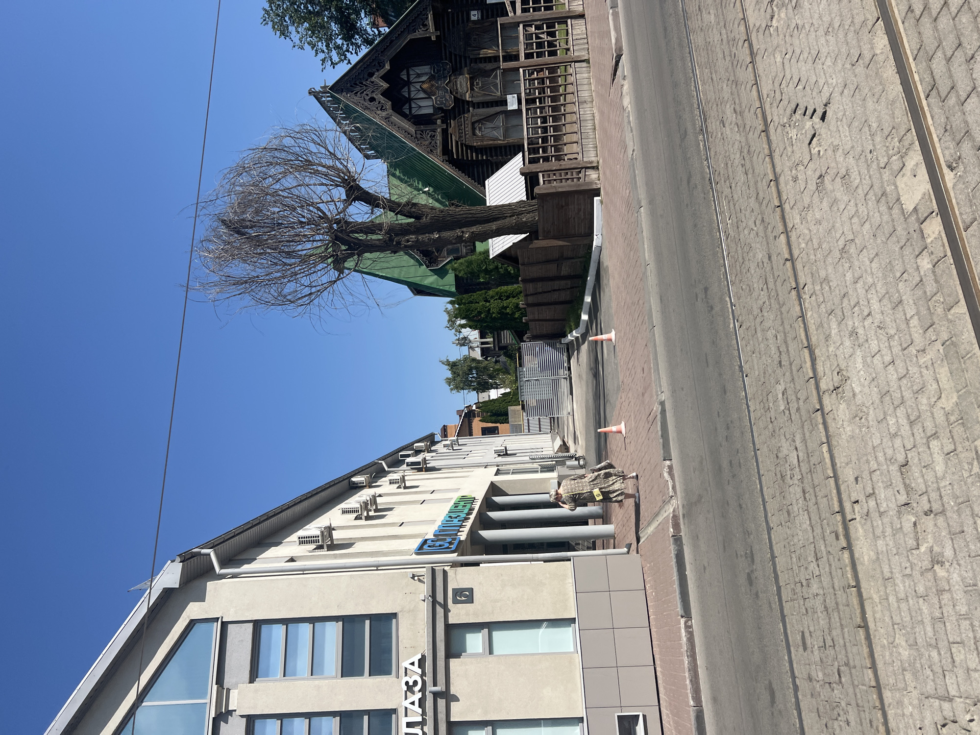 A street view in Ulyanovsk, Russia shows a modern office building on the left and a traditional wooden house on the right. The office building has a light-colored facade with large windows and an angled roofline. Air conditioning units are visible on its exterior. A woman in a patterned dress is walking away from the camera on the sidewalk in front of the building. On the right, a dark wooden house with intricate carvings and a green roof stands behind a wooden fence. A bare tree with thin branches is positioned in front of the wooden house. The street in the foreground is paved with bricks, and tram tracks are embedded in the pavement. The sky is clear and blue. Orange traffic cones are placed on the sidewalk.