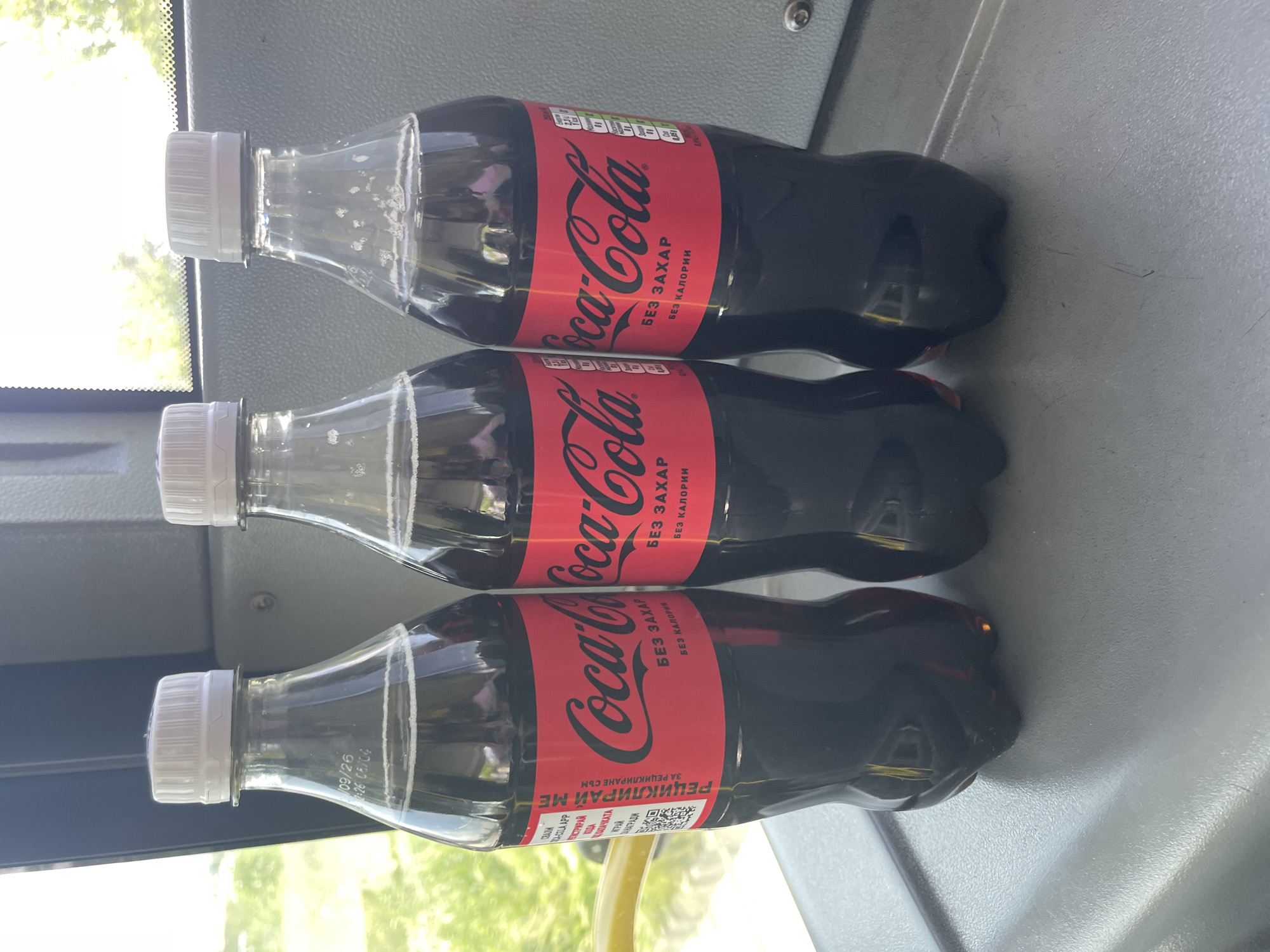 Three bottles of Coca-Cola Zero are arranged horizontally on a gray surface, possibly a seat on public transportation. The labels are red with "Coca-Cola" printed in the classic white font, along with "БЕЗ ЗАХАР БЕЗ КАЛОРИИ" which translates to "No Sugar No Calories" in Bulgarian. The bottle closest to the bottom of the image has a recycling message "РЕЦИКЛИРАЙ МЕ ЗА РЕЦИКЛИРАНЕ СЪМ" translating to "Recycle Me I'm for recycling" along with a QR code. The bottles are capped with white tops. The background shows a window with greenery visible. The labels appear to be in Bulgarian. The presence of Cyrillic text strongly suggests the location is Plovdiv, Bulgaria. The location is most likely a bus given the seat surface, window, and objects in the background.
