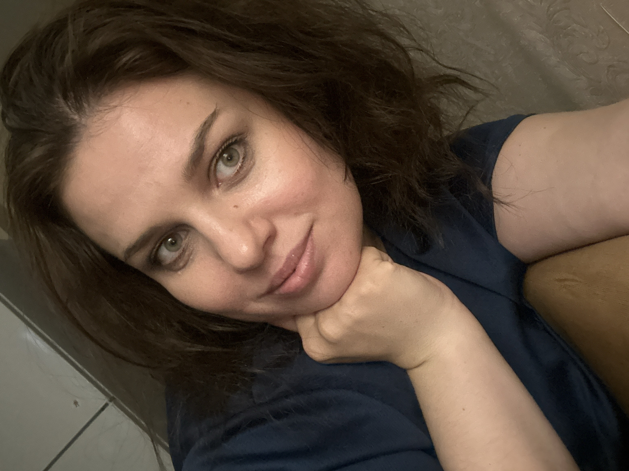 The image shows a close-up, selfie-style photograph of a woman's face and upper torso. She has medium-toned skin, dark brown hair that is slightly disheveled, and light green eyes. Her left hand is resting on her chin, and she is wearing a dark blue t-shirt. The background appears to be indoors, with textured walls and what looks like a tiled floor or surface in the upper left corner. The lighting is dim, suggesting the photo was taken in low-light conditions.