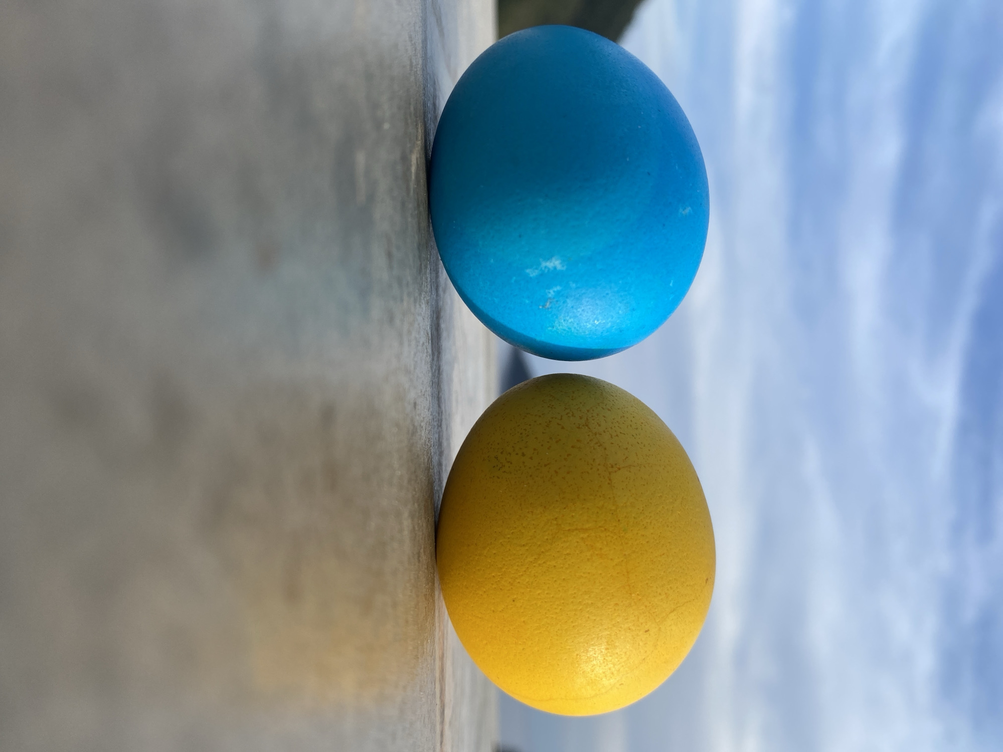 Two painted eggs, one blue and one yellow, are stacked vertically on a concrete surface. The eggs' coloring appears to be slightly textured. The background consists of a cloudy sky. The scene is likely located in Budva, Montenegro, given the colors of the eggs mirroring the flag of Ukraine.