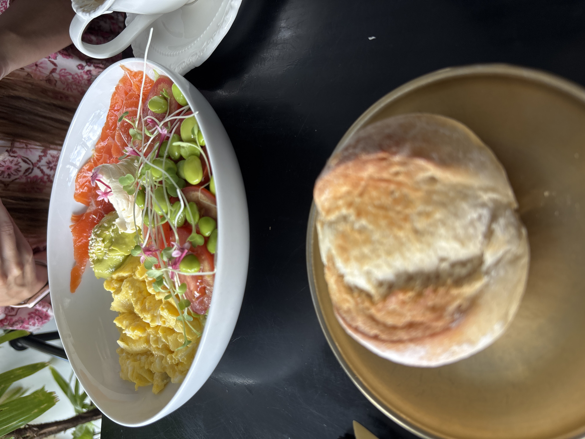 A bright, overhead shot captures a delectable brunch spread on a dark, reflective table, likely in Kuta Selatan, Indonesia. The central focus is a large white bowl overflowing with a colorful and textured assortment of food. Scrambled eggs, a vibrant yellow, form a substantial base. Nestled beside them are thinly sliced pieces of smoked salmon, their rich orange hue a striking contrast. A dollop of creamy white sauce, possibly cream cheese or a similar spread, is garnished with delicate pink flowers and microgreens. Scattered throughout the bowl are bright green edamame beans and chopped red tomatoes, adding pops of color and freshness. A portion of sliced avocado, a subtle green, is visible near the salmon, lightly sprinkled with sesame seeds.
In the foreground, slightly out of focus, a golden-brown bread roll rests on a round, gold plate. The roll has a rustic appearance, with a well-risen top and a slightly cracked crust. To the right of the main bowl, a white ceramic pitcher with a handle sits on a white saucer, suggesting the presence of a beverage, perhaps coffee or tea. A sliver of a person's arm, clad in a pink floral-patterned garment, is visible in the upper right quadrant, indicating someone is present and likely enjoying the meal. The overall impression is one of a luxurious and satisfying breakfast or brunch experience in a well-lit, possibly indoor setting with lush green foliage visible in the upper left corner.