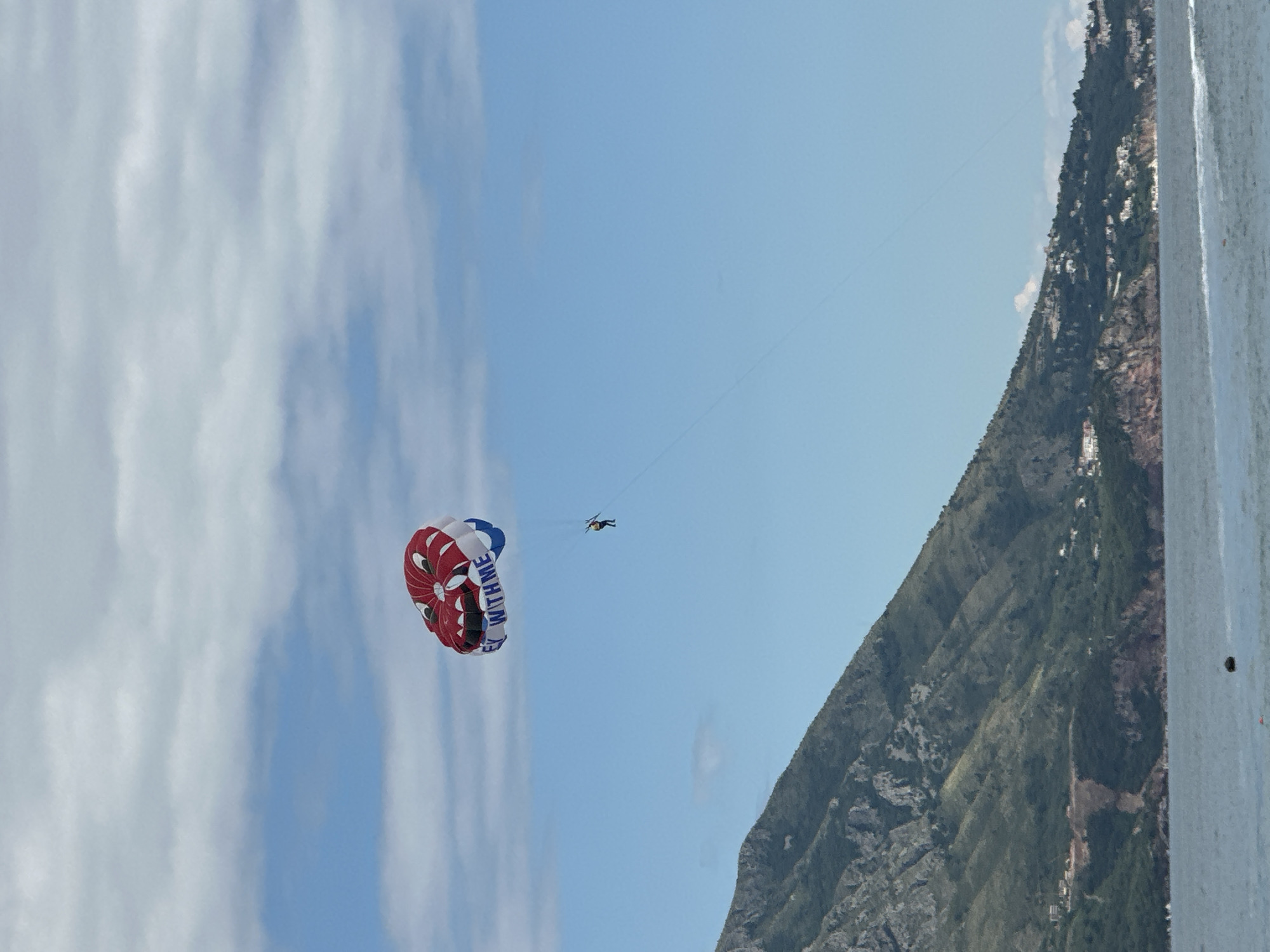 The image shows a person parasailing over a mountainous coastal landscape under a clear blue sky. A brightly colored parasail, primarily red and white with a blue section, is prominent in the upper left portion of the frame. The words "FLY WITH ME" are visible in white text on the blue part of the parasail. Suspended beneath the parasail, a single individual is seen, appearing small in the vast sky, indicating they are high above the ground or water.
To the right, a large, verdant mountain or hillside dominates the background, sloping downwards towards the bottom right. The mountain is covered in dense green foliage and features several small buildings and structures scattered across its slopes. The sky, which occupies the majority of the upper frame, is a vibrant blue with sparse, wispy white clouds. A thin line, likely the tow rope for the parasail, extends from the parasail towards the right, leading out of the frame. A small, indistinct part of a boat or platform is visible along the extreme right edge of the image. The scene suggests a bright, sunny daytime setting, characteristic of good weather for parasailing in a coastal region like Budva, Montenegro.