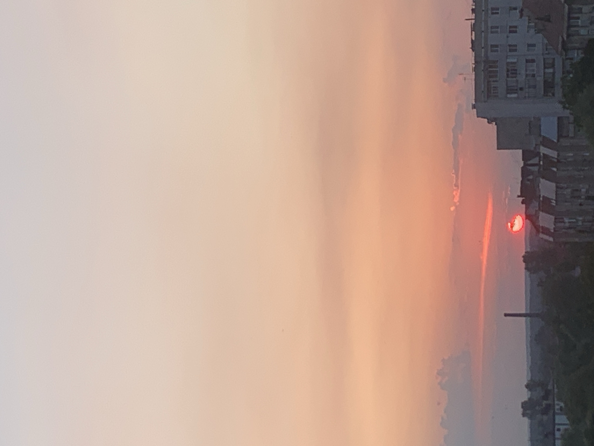 The image shows a sunset over the city of Kolomyia, Ukraine. The sky is a gradient of pink and orange, with a bright red sun setting on the horizon. In the foreground, there are buildings and trees, with a tall smokestack visible in the distance. The buildings appear to be apartment blocks, with some houses visible as well. The trees are lush and green, indicating that it is likely summer. The overall impression is one of a peaceful, urban landscape bathed in the warm glow of the setting sun.