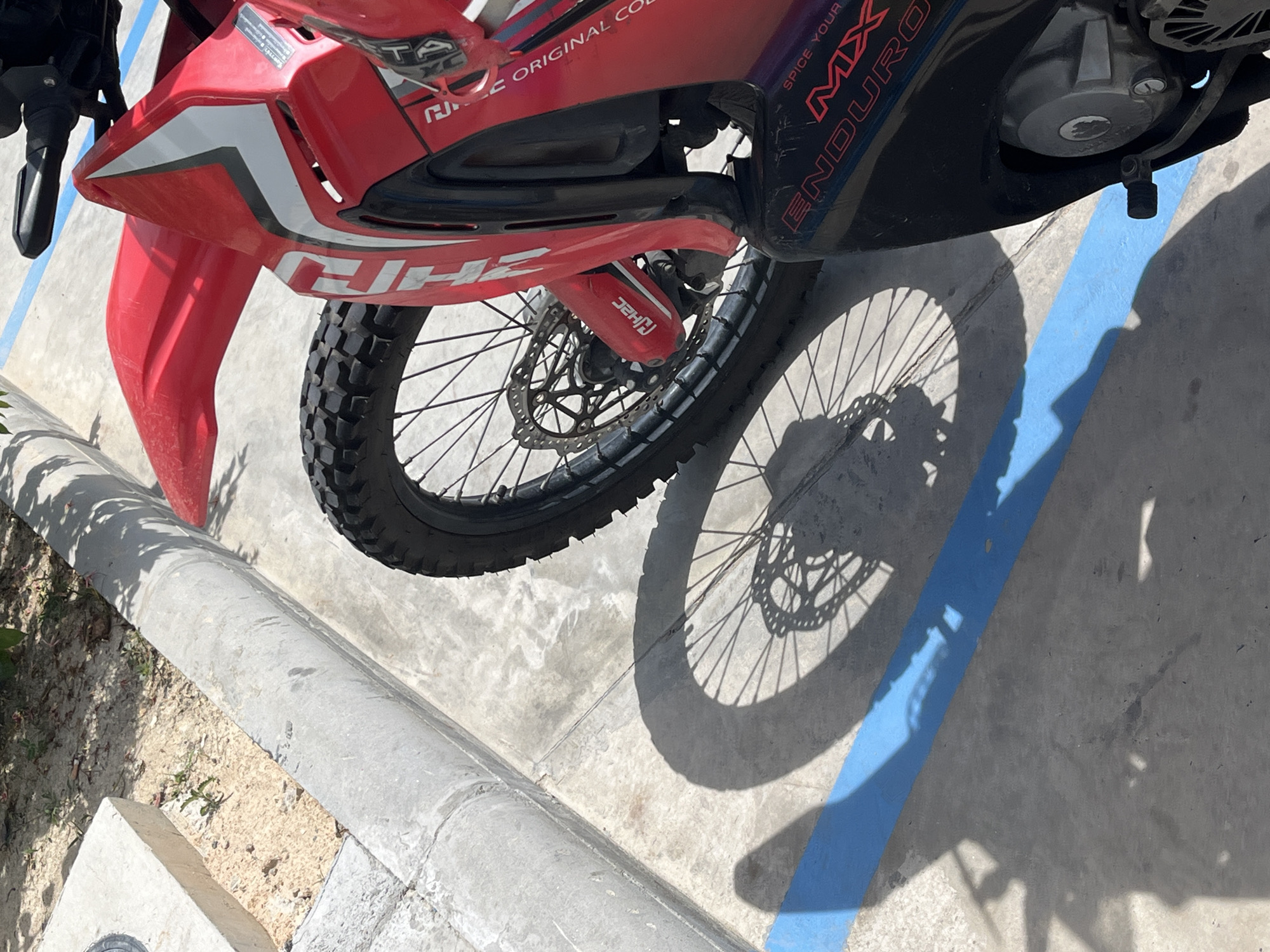 A red and black dirt bike is parked on a concrete surface next to a curb in a city environment. The motorcycle has knobby tires, a red fender, and visible branding that includes "ENDURO" and "MX". A shadow of the motorcycle's wheel and spokes is cast across the concrete and a bright blue line painted on the surface. The background includes sandy soil, sparse vegetation, and a concrete structure. The lighting suggests it is daytime.
