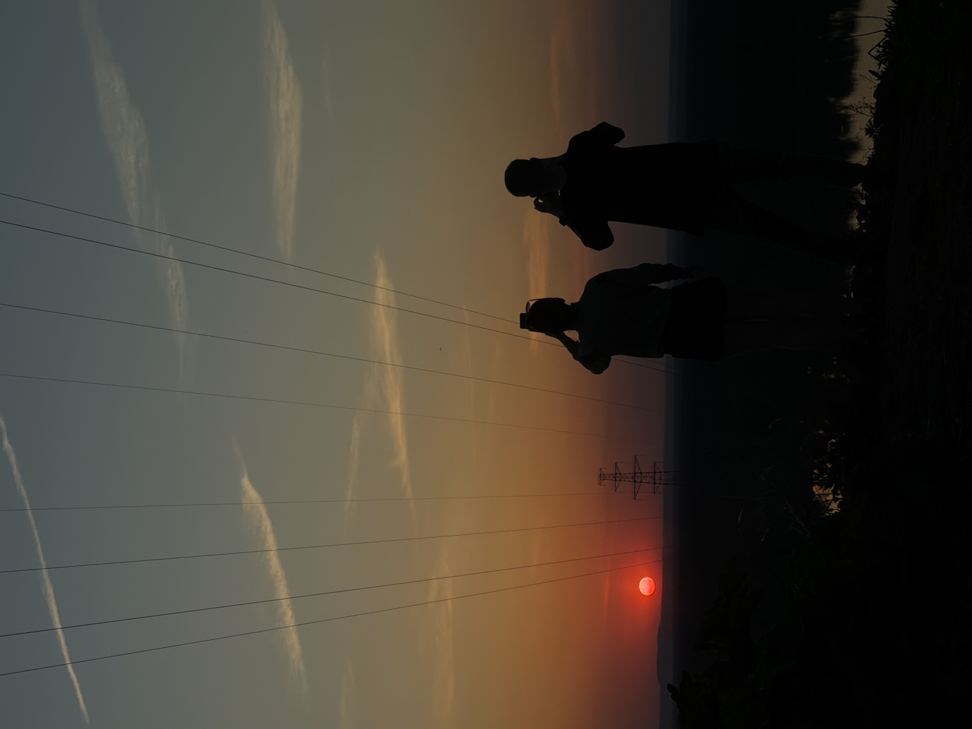 The image captures a sunset scene in Noviy, Russia. Two silhouetted figures stand in the foreground, presumably taking photographs of the sunset with their mobile phones. Overhead power lines stretch across the sky, leading to a lattice steel transmission tower in the distance. The sun appears as a bright red orb on the horizon, casting a warm orange hue across the sky. The ground beneath the figures is dark and indistinct, likely vegetation. There is what appears to be some streaky cirrus clouds in the sky.