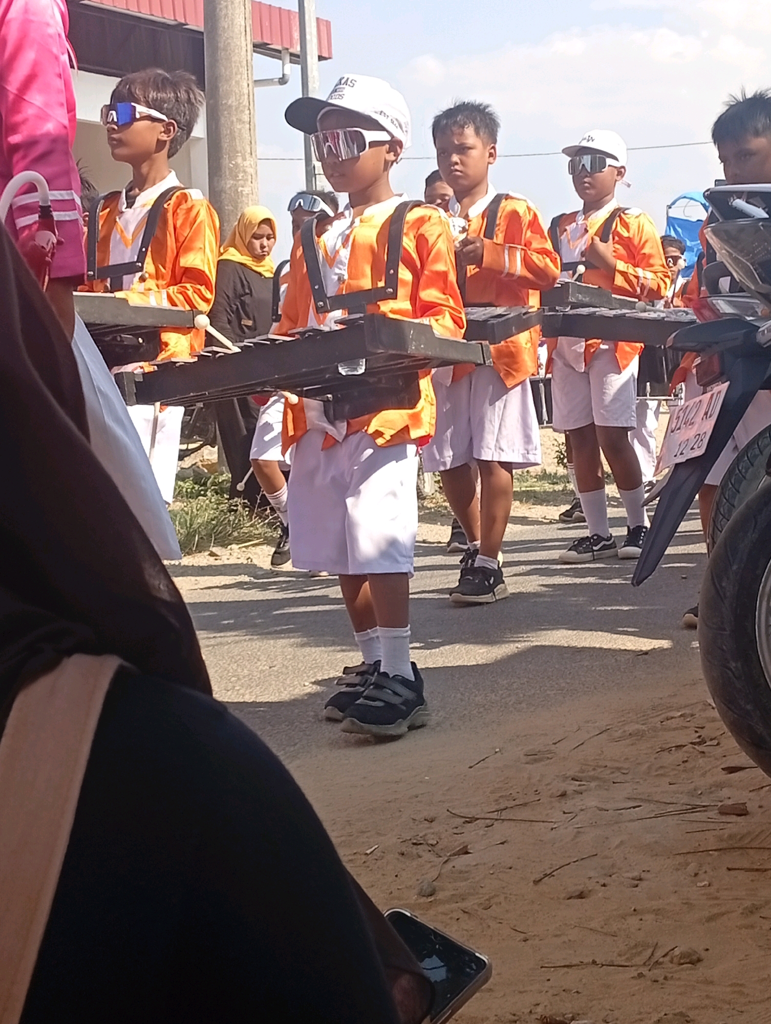 The image depicts a street scene in Nglojo, Indonesia, during what appears to be a parade or performance. Several young boys, dressed in matching orange and white marching band uniforms, are positioned in a line. They are wearing white hats or caps and distinctive white-rimmed goggles. Each boy is holding a set of mallets, suggesting they are about to play percussion instruments, possibly xylophones or glockenspiels, which are visible in front of them. The uniforms include orange jackets with white accents and white shorts or skirts, paired with white socks and black sneakers. The ground is a mix of asphalt and sandy dirt, with some dry leaves and twigs scattered around. In the background, a woman wearing a yellow hijab is visible, as are other figures and what appears to be a motorcycle on the right side of the frame. The overall atmosphere is one of organized activity under a bright, possibly sunny sky.