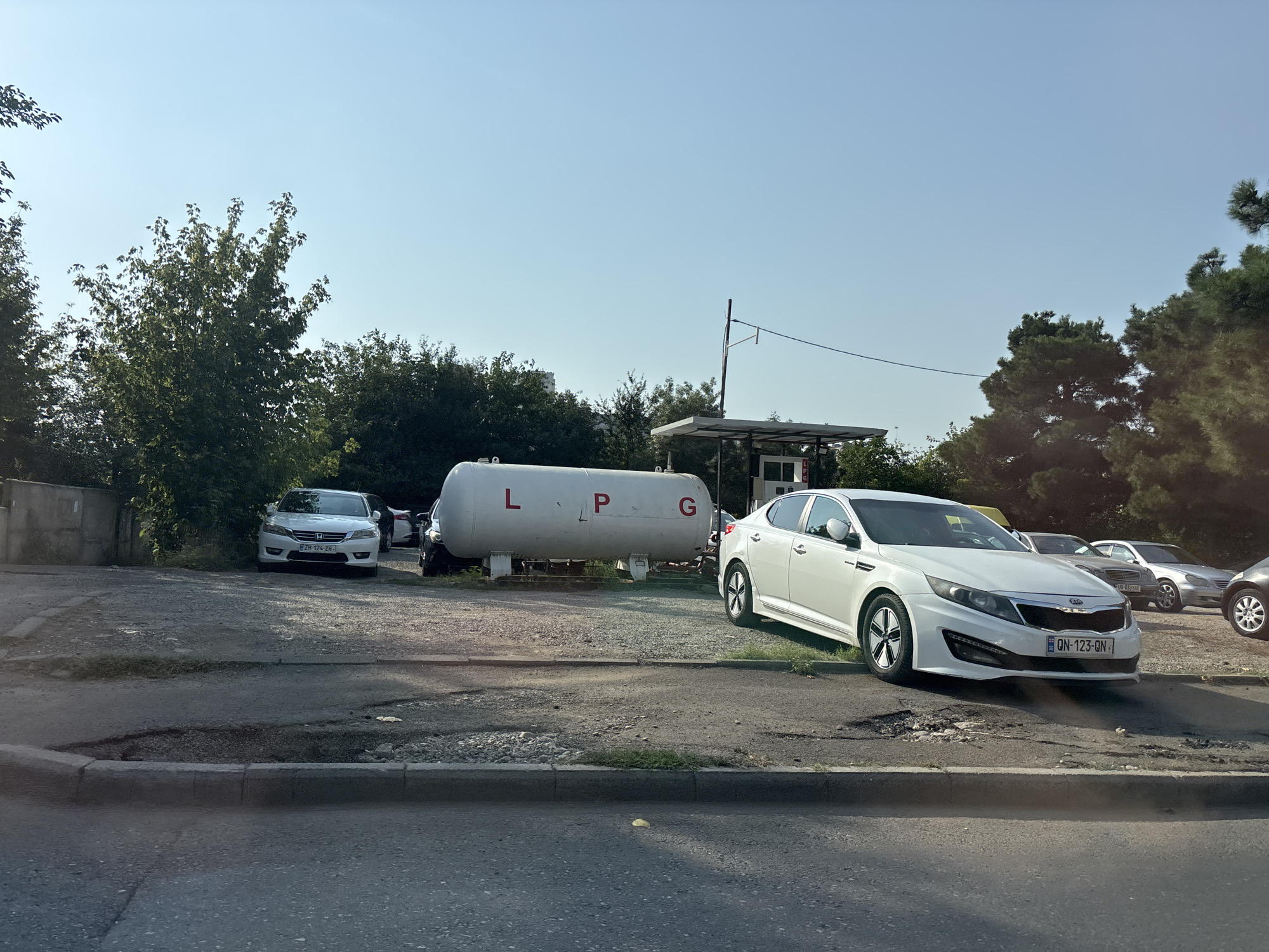 The image depicts an outdoor scene in Tbilisi, Georgia, likely a gas station or depot. A large white cylindrical tank with "LPG" in red letters is centrally located, suggesting it stores liquefied petroleum gas. Several cars are parked around the tank and in the background, including a white Kia Optima in the foreground, displaying the license plate GN-123-ON. To the left, a white Honda Accord with license plate ZH-174-ZH is parked. Behind the LPG tank, a small covered structure with a sign that appears to read "LPG" is visible. The ground is a mixture of gravel and asphalt, with some patches of vegetation. Trees line the background, and the sky is clear and blue. The overall impression is one of an operational industrial or commercial area.