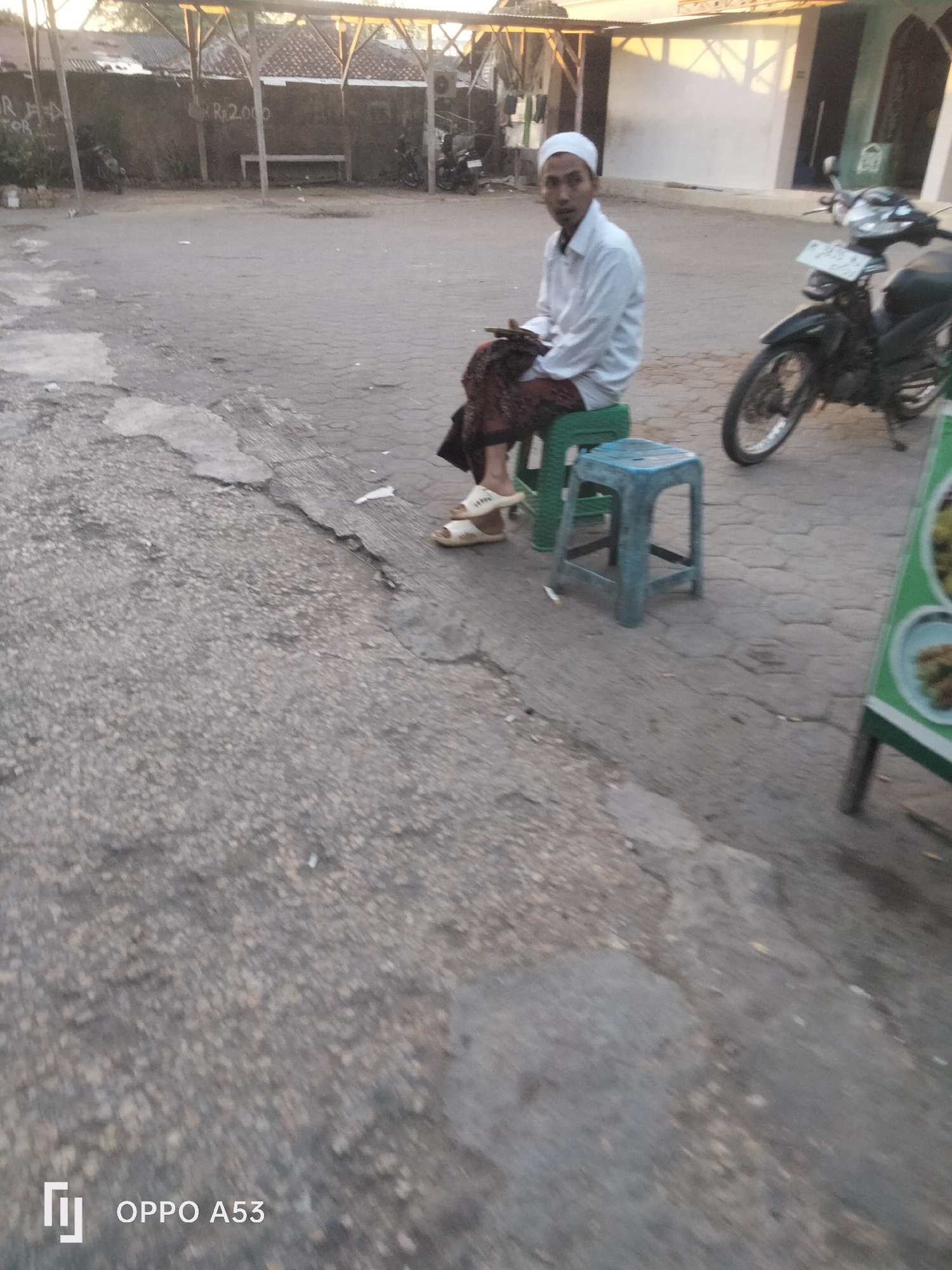The image depicts a street scene, likely in Badung, Indonesia, characterized by a gray, cracked asphalt surface. In the mid-ground, a man wearing a white shirt and peci (a traditional cap) is seated on a green plastic stool. He is wearing a patterned sarong and white sandals. Next to him is a smaller blue plastic stool. To the right of the man, a dark-colored motorcycle is parked. In the background, there is a low wall with some graffiti, including "Rp 2.000". A covered structure with wooden supports is visible behind the wall, and buildings with white walls and windows are further in the distance. A green menu board is partially visible on the far right of the frame. The lighting suggests it is either early morning or late afternoon.