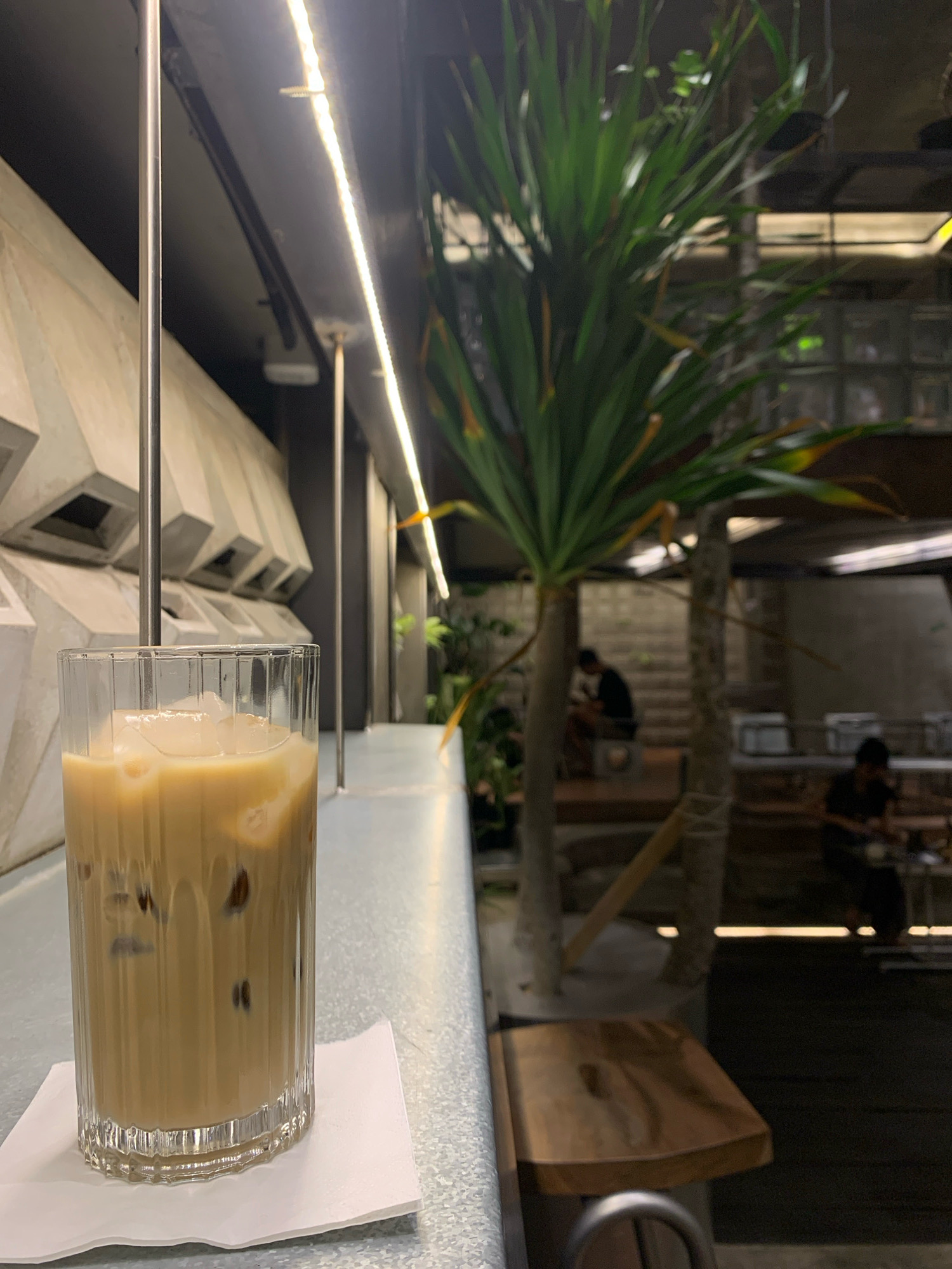 The image shows an iced coffee in a fluted glass on a metal counter in a cafe. The drink is light brown with ice cubes. In the background, there is a large plant, and further back, there are blurred figures of people sitting at tables in a dimly lit indoor/outdoor space. The architectural style of the cafe appears modern with concrete elements and exposed lighting. The overall ambiance suggests a contemporary coffee shop. Without further information, the specific location in Mojokerto, Indonesia, cannot be determined from the visual elements alone.