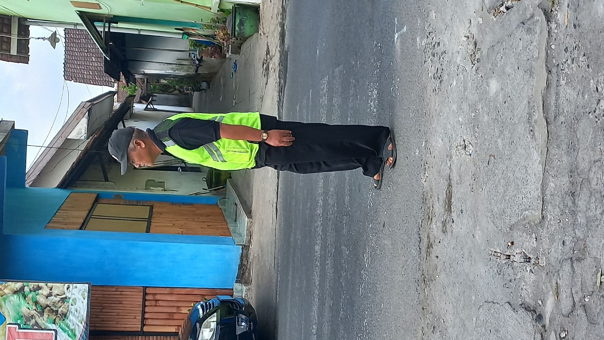 A man wearing a black shirt, black pants, a black baseball cap, and a neon green safety vest stands facing right on a paved street. He is wearing dark sandals. The street is cracked and shows signs of wear. To his left is a dark-colored car. Behind the man, a narrow alleyway runs between two buildings. The building on the left is painted bright blue with wooden accents. The building on the right is painted green. The alleyway has various objects, including potted plants and a hanging light fixture. The scene appears to be in a residential or commercial area in Kenayan, Indonesia.
