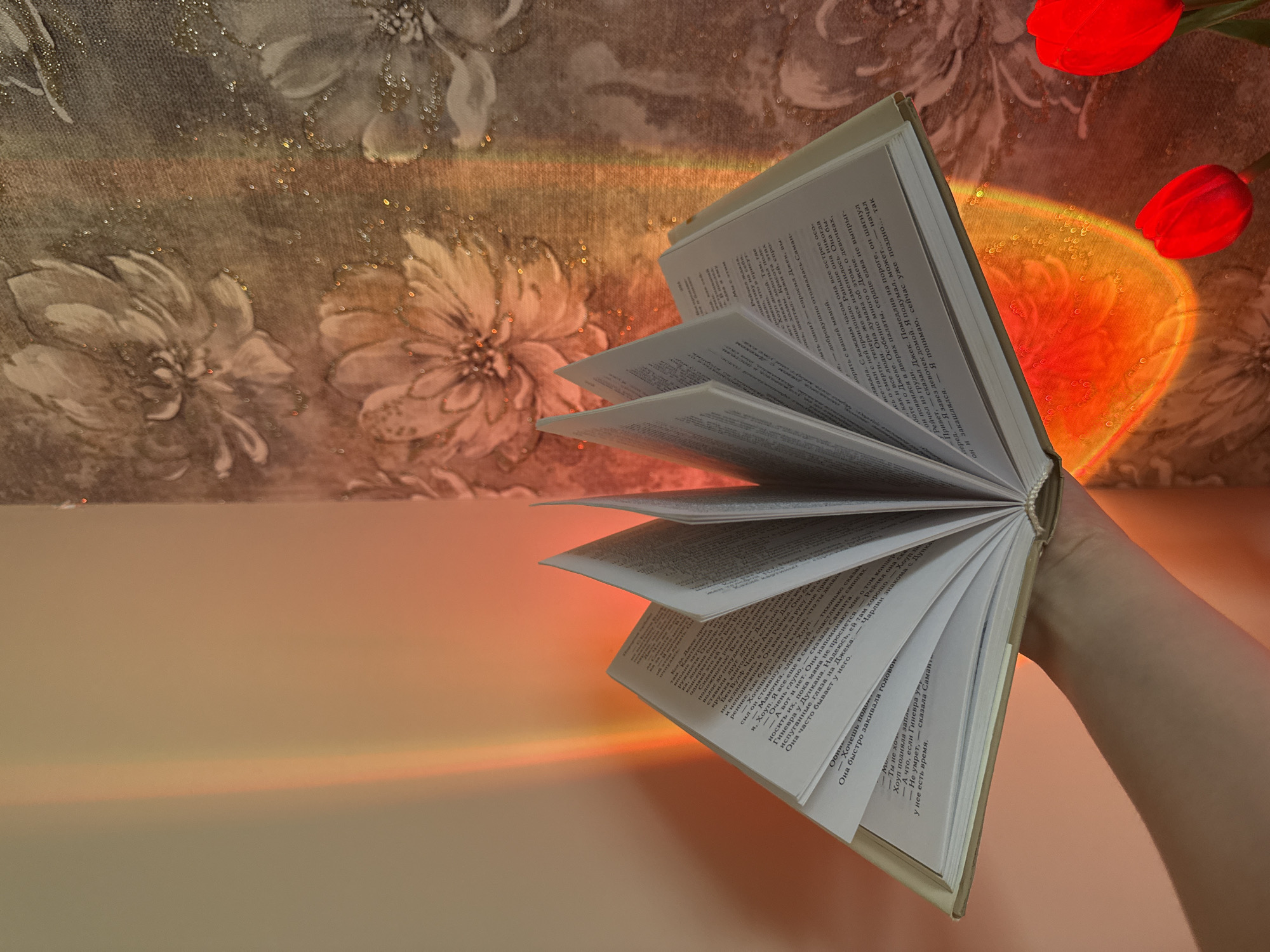 The image shows an open book held by a person's hand, with pages fanned out. The book is illuminated by a warm light, possibly from a projector, creating colorful gradients and reflections on the wall behind it. The wall has a floral pattern and sparkles. Two red tulips are visible in the lower right corner, partially obscured by the light. The scene is indoors, likely a domestic setting.