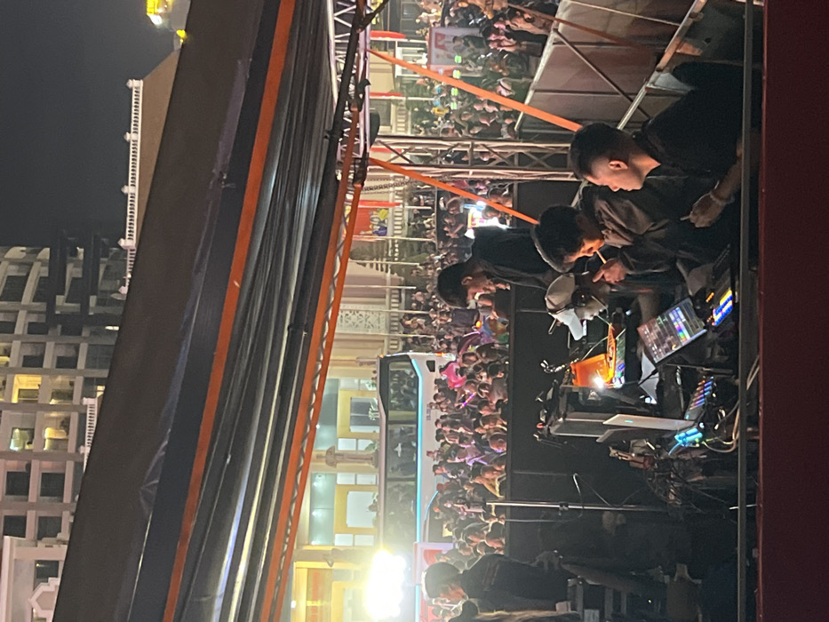 The image depicts an outdoor night scene in Nha Trang, Vietnam, specifically in the vicinity of the Nha Trang Cathedral, identifiable by its Gothic architecture, prominent archways, and a visible statue in the background.
The foreground is dominated by a temporary stage or control platform covered by a black and orange canopy structure. Four males are present on this platform. On the far left, a male is partially visible, standing. To his right, another male in a black shirt stands, looking downwards. In the center-right, two males are seated at a control station equipped with multiple laptops displaying illuminated screens, audio or lighting mixing consoles with glowing buttons, and various cables. The seated male on the left wears a grey jacket and holds a lit cigarette to his mouth while looking at the equipment. The seated male on the right wears a black shirt and holds a thin white object, consistent with a cigarette or vape pen, to his mouth, looking towards the right. A white baseball cap and an orange lamp are also visible on the control station. Metal trussing supports the overhead structure.
Behind the platform, a large crowd of people is densely gathered, facing forward, suggesting an audience at an event. Some individuals in the crowd are holding illuminated objects. A white tour bus or large passenger vehicle is visible in the mid-ground, positioned within or behind the crowd. The background features the illuminated Nha Trang Cathedral and, further back, a modern, multi-story building with multiple lit windows, contrasting against the dark night sky. The scene indicates a public gathering or performance under artificial lighting.