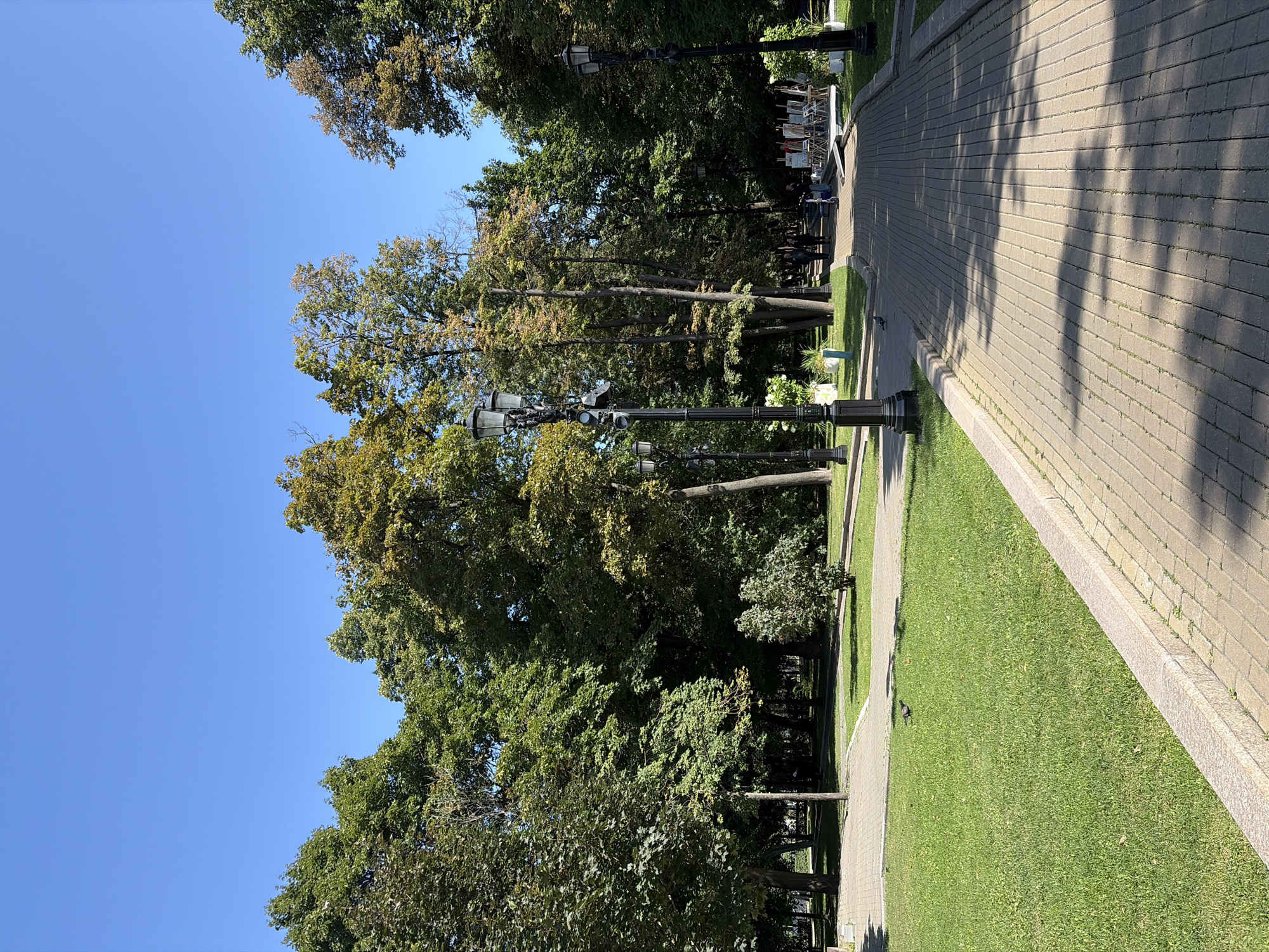 This is an eye-level, full-shot photograph of a paved park pathway in Moscow, Russia, under a clear blue sky on a sunny day. The pathway is made of light brown rectangular bricks and curves slightly to the right, leading into the distance. The path is lined on the left by a well-maintained, vibrant green lawn with scattered fallen leaves. Beyond the lawn, a dense line of mature trees with lush green foliage forms a canopy, with some trees showing hints of yellow and brown, suggesting the early onset of autumn. On the right side of the pathway, there is more green grass and a continuation of the tree line.
Along the pathway, several black, ornate lampposts are visible. The closest lamppost on the left is in the foreground, with three lamps and decorative detailing. Further down the path, two more lampposts are visible, one on the left and one on the right.
In the middle ground, a few people are walking along the pathway. Two individuals are walking together, and two more are ahead, one of whom is pushing an electric scooter. Further into the park, a small art exhibition is set up with easels displaying artwork and tables. White planters with greenery and white flowers are placed near the lampposts. Two pigeons are visible on the grassy area to the left of the pathway. The overall impression is a peaceful and well-maintained urban park.