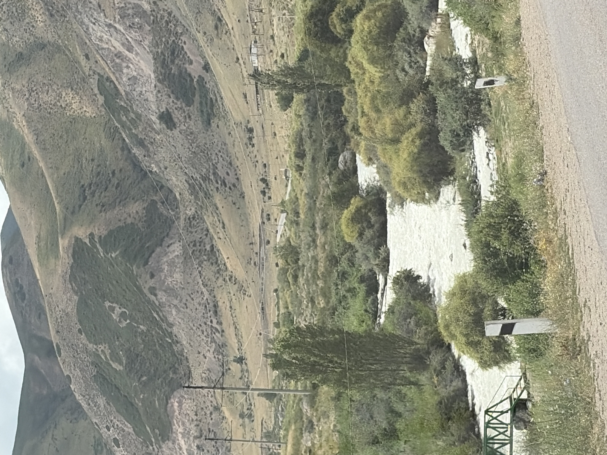 The image depicts a natural landscape with a river flowing through a valley. In the foreground, a portion of an asphalt road is visible, with a green metal bridge crossing the river on the left. A white marker post stands beside the road, partially obscured by vegetation. The river itself is wide and appears to be flowing rapidly. Dense green bushes and trees line the riverbanks and extend into the valley. In the background, rolling hills rise up, covered in dry grass and sparse vegetation, with some areas appearing rocky or eroded. Power lines and utility poles are also visible in the middle ground, suggesting the presence of infrastructure. The sky is overcast, casting a diffused light over the scene. The overall impression is one of a rural or undeveloped area, possibly in a mountainous region.