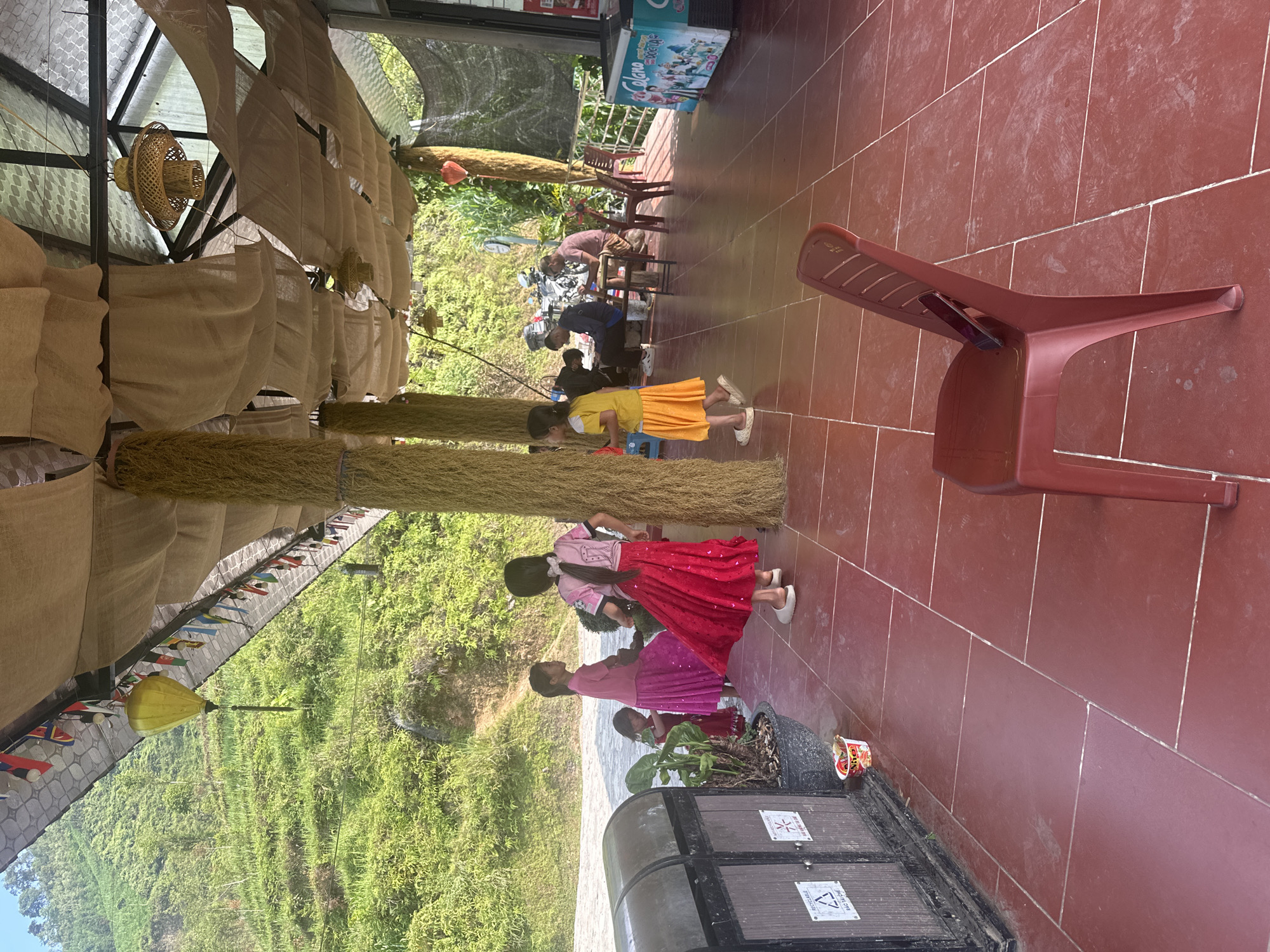 Scene in Đường Thượng Commune, Vietnam, showing a covered patio area with a red tiled floor. Three children are present: one in a yellow dress and two in pink tops with red and purple skirts. A red plastic chair sits on the patio. Additional people are visible in the background seated at a table. The patio is adorned with decorations including straw elements and fabric. In the foreground, there is a black container holding a potted plant. Beyond the patio, there is a view of a green hillside. A cooler and hanging lights are also visible.