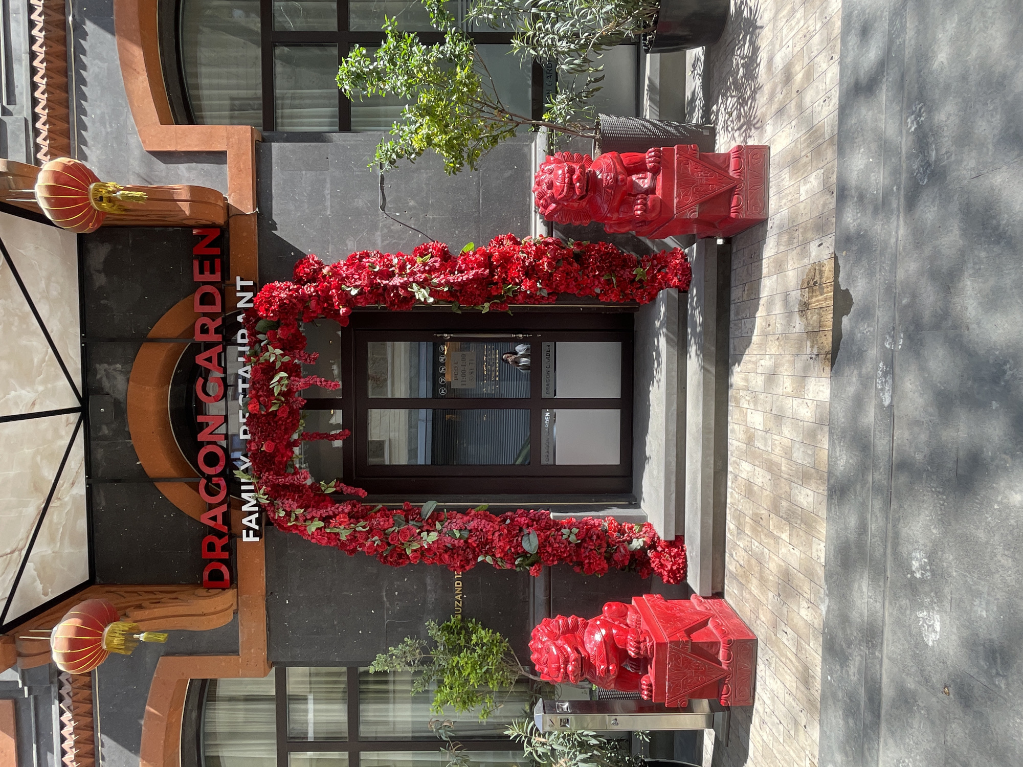 The image shows the exterior of the "Dragon Garden Family Restaurant" located at Suzand 12 in Yerevan, Armenia. The restaurant's name is prominently displayed in red lettering, with the words "Family Restaurant" below. A red floral arrangement arches over the entrance, which is flanked by two bright red, traditional Chinese lion statues, or foo dogs, on small platforms. The building facade features a dark gray finish with brown and red trim. A paper lantern hangs on the left side of the entrance, adding to the oriental ambiance. The sidewalk in front of the entrance is made of light-colored stone or brick. The restaurant appears to be open for business as signage is visible on the doors.