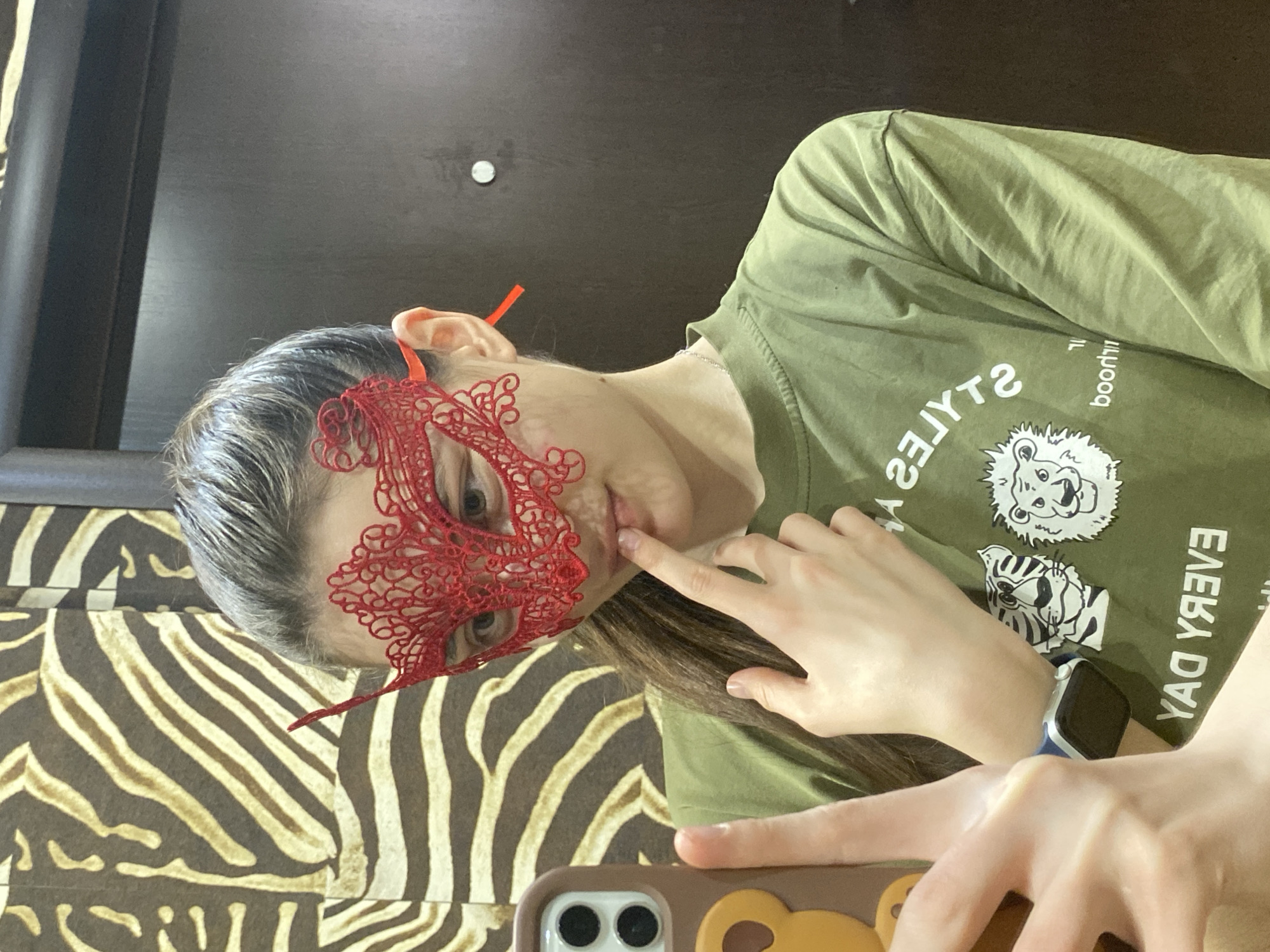 An indoor shot features a person, likely female, captured from a low angle. The person is wearing a striking red, intricately patterned lace mask that covers their eyes and part of their nose. Their hair appears dark brown or streaked with grey, parted on the side. They are wearing an olive green t-shirt with mirrored white text and graphics. The text, when viewed in a mirror or corrected, reads "STYLES" and "good" and "EVERY DAY," accompanied by drawings of a lion's head and another feline, possibly a tiger.
The person's right index finger is gently pressed against their lips, conveying a pensive or secretive expression, while their eyes, visible through the mask, look towards the viewer. A smartwatch with a blue band and a dark screen is visible on their left wrist.
The background features a prominent brown and light yellow/beige zebra-stripe pattern on a wall, suggesting patterned wallpaper or fabric. Above the person's head is a dark brown, possibly wooden, surface. In the immediate foreground, the top portion of a smartphone is visible, encased in a brown cover featuring a cartoon bear design and showing the camera lenses. The overall scene suggests a self-portrait or mirror selfie, possibly for a playful or mysterious mood. The lighting is internal and soft, with no specific time of day indicated. There are no direct cues to the provided location of Votkinsk, Russia.