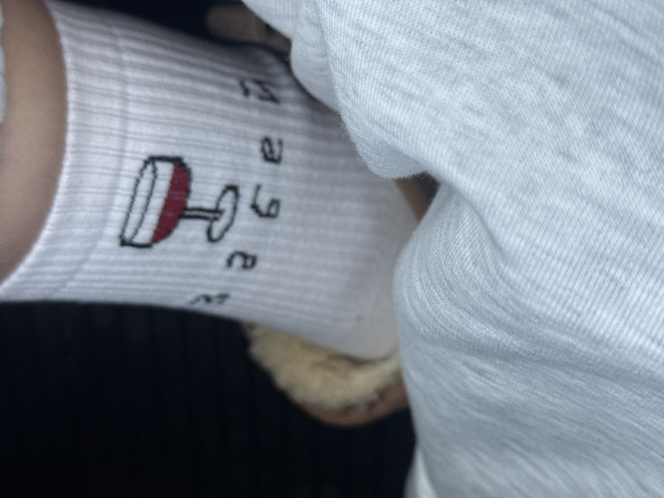 A close-up image displays a lower leg or foot, partially obscured, wearing a white ribbed sock. The sock features a graphic of a wine glass filled with red liquid, accompanied by Cyrillic text which reads "ЗАБЕЙ". The foot appears to be resting in a light-colored, possibly faux-fur lined slipper, partially visible behind the sock. The background is dark and out of focus, suggesting an indoor setting with limited illumination. A white, textured fabric, likely clothing or bedding, occupies the lower foreground. The specific location within Achkhoti, Georgia, cannot be determined from the visual information provided.