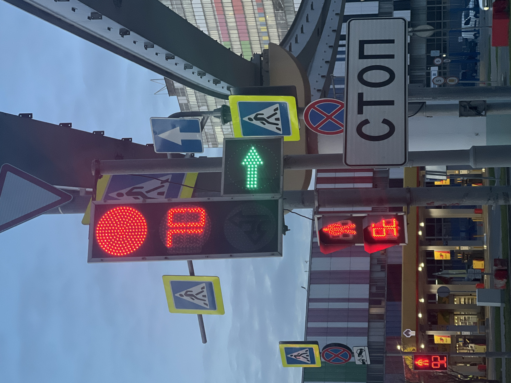 The image captures a busy urban street scene in Khimki, Russia, filled with numerous traffic signals and road signs under an elevated structure or overpass. The sky is overcast and uniformly grey, suggesting a dull day or possibly late afternoon/early evening given some illuminated lights in the background buildings.
Dominating the center-left, a large traffic light unit displays a prominent red circle and a red stylized 'P' symbol (Cyrillic 'R'), which in Russia commonly indicates 'Don't Walk' for pedestrians. Adjacent to this, a smaller, square traffic light illuminates a bright green arrow pointing straight, signaling that straight-moving vehicular traffic is permitted.
Above and to the right of these lights, a tall white rectangular sign is visible with clear black Cyrillic text reading "СТОП" (STOP). Nearby, a round red sign with a diagonal line through it indicates 'No Stopping' or 'No Parking'. Several blue square signs with white pedestrian figures inside a yellow border, marking pedestrian crossings, are also scattered throughout the frame. One such sign is prominently displayed above the green arrow light, another below the main traffic light, and a third further right.
On the right side of the image, two more red traffic lights are active. One shows a red pedestrian figure along with the number '84' in red, likely a countdown timer for pedestrians waiting to cross. The other displays a red left-pointing arrow, also accompanied by the '84' countdown, presumably for vehicles intending to turn left. A blue square sign with a white right-pointing arrow is also present, directing traffic.
The background reveals modern buildings with multiple windows, some reflecting light, and the robust metal structure of the overpass with a patterned roof, possibly glass. The combination of numerous signals and signs highlights a complex intersection designed for efficient traffic and pedestrian management.