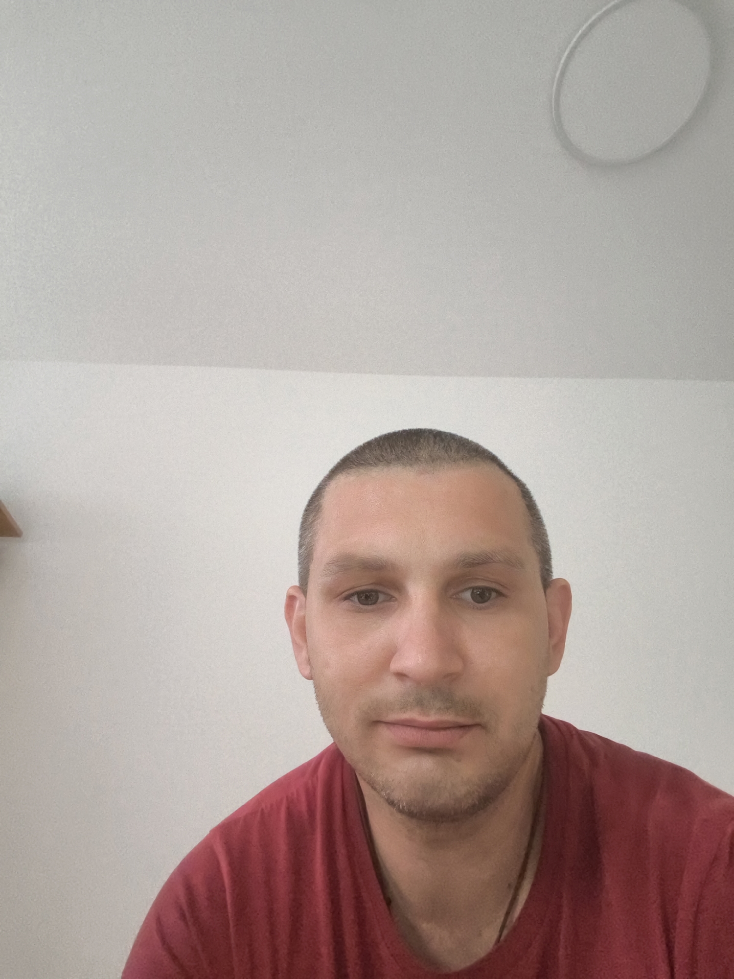 A close-up, waist-up selfie of a white man with a buzzed haircut and light stubble is taken from a slightly low angle. He is wearing a dark red t-shirt. The background consists of a white wall with a faint circular light fixture on the ceiling. The image appears to be taken indoors, likely in a residential setting. There is a shelf visible on the left side of the frame. The location in Wolgast, Germany is not discernible from the image.