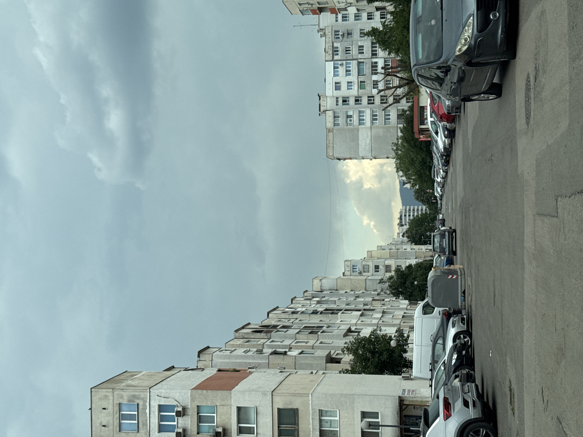 The image depicts a street scene with apartment buildings and parked cars, likely in Sofia, Bulgaria. The buildings are multi-story, utilitarian in style, and appear to be a mix of older and more modern construction. Some buildings exhibit a tiered or stepped architectural design, creating an interesting visual pattern. Numerous cars are parked along the side of the street, indicating a residential area.
The sky is overcast with dramatic clouds, suggesting a cloudy or possibly stormy day. The lighting is diffused, typical of an overcast sky, and there are no strong shadows. The time of day is not clearly discernible but appears to be daytime. The overall mood is somewhat somber due to the weather conditions.
There is no visible text in the image. The focus is on the urban landscape, the architecture of the apartment blocks, and the line of parked vehicles. The image has been rotated 90 degrees clockwise, so the buildings appear to be standing on their sides.