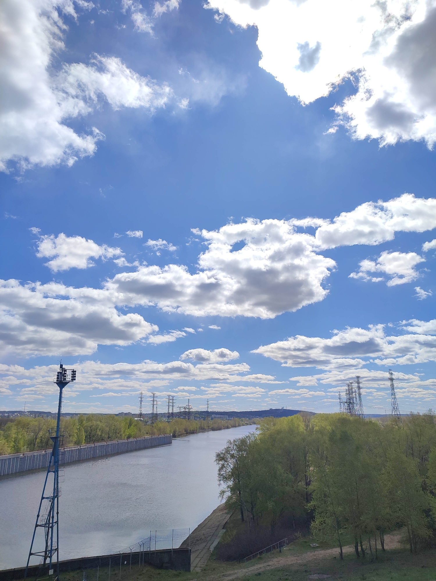 A sunny, daytime shot of a river canal with a steel retaining wall on one side, a grassy bank with trees on the other, and an industrial area in the background. The sky is a vivid blue with scattered white cumulus clouds. A tall metal tower with floodlights stands on the side of the canal. In the distance are power lines and towers. The location is in the city of Unknown, Unknown.