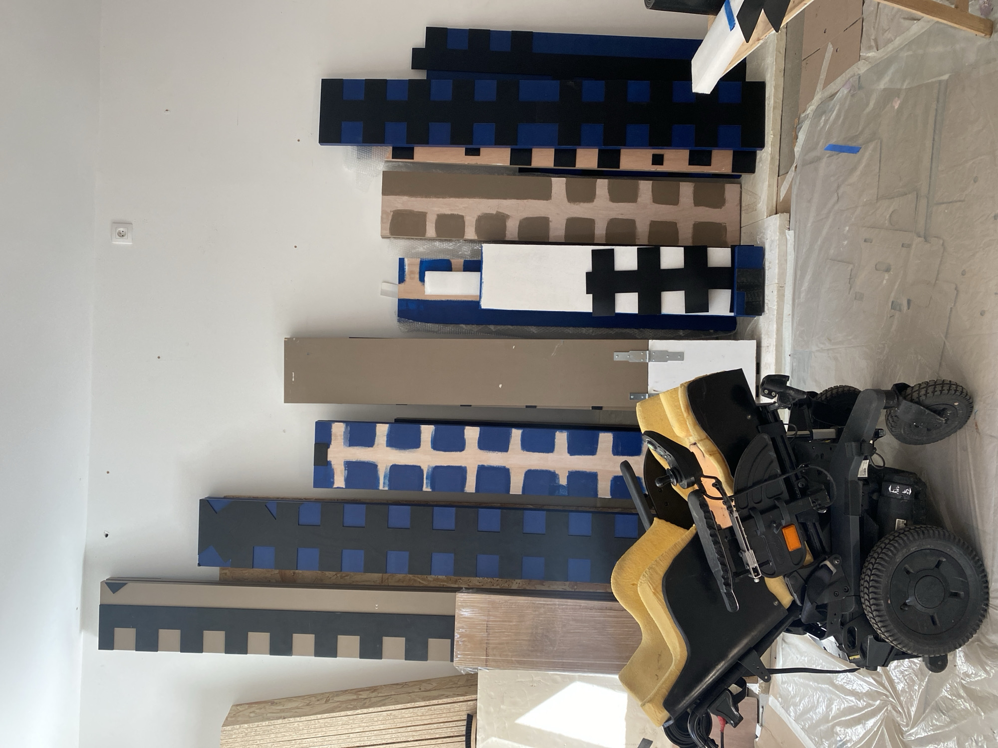 A brightly lit indoor scene captures a collection of wooden planks, some plain and some decoratively painted, leaning against a white wall. The painted boards display various geometric patterns, including black and blue squares, dark brown and beige squares, and black and white squares. Other designs feature blue squares on a light brown background, dark blue segments on a lighter base, and grey and beige square patterns. Some boards also incorporate triangles and an 'H' shape.
In the foreground on the right, a black electric wheelchair with prominent yellow/tan upholstered cushions is visible. It appears to be in a compact or partially folded state, resting on a sheet of clear plastic covering the floor. The floor itself is mostly covered by this plastic sheeting, with blue painter's tape visible in places, suggesting ongoing painting or renovation work. Stacks of unpainted wooden boards are also visible towards the bottom left of the image.
The white wall features a single, standard European-style power outlet. The overall impression is a workshop or a room undergoing renovation in Paris, France, with a focus on crafting or preparing decorative panels. No legible text is visible on any of the objects.