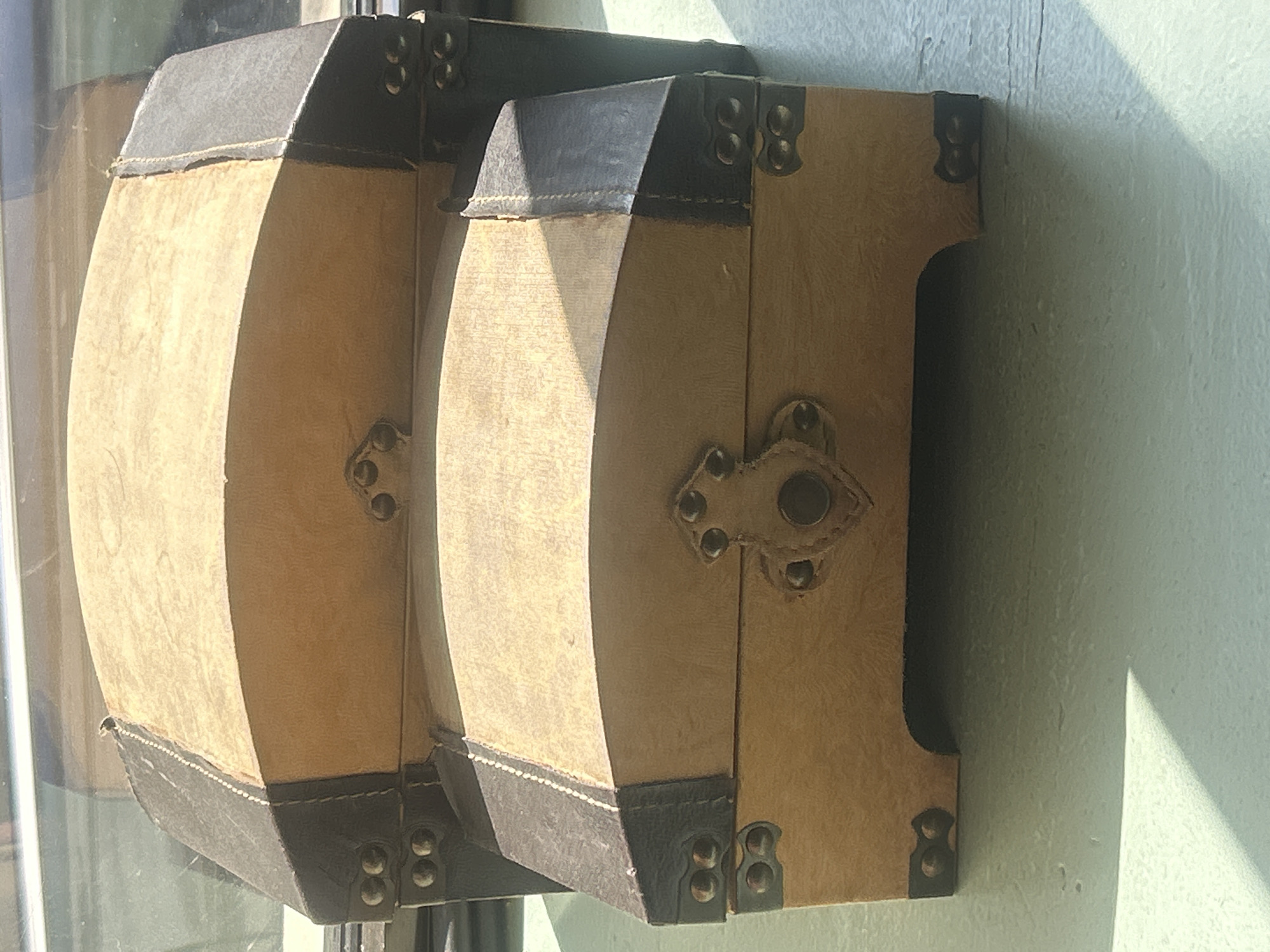Two vintage, faux-leather-covered wooden chests are stacked on a light-colored, textured surface. The chests are light brown with dark brown trim and decorative metal studs. The top chest is larger and has a curved lid, while the bottom chest is smaller and also has a curved lid. Both chests have ornate metal clasps in the center of their lids. The scene appears to be indoors, near a window, as evidenced by the sunlight casting shadows and reflections. The background shows a blurred window and possibly a ledge. There are no people or animals present. The image is cropped to focus on the chests. There is no information to suggest the location is Almaty, Kazakhstan.