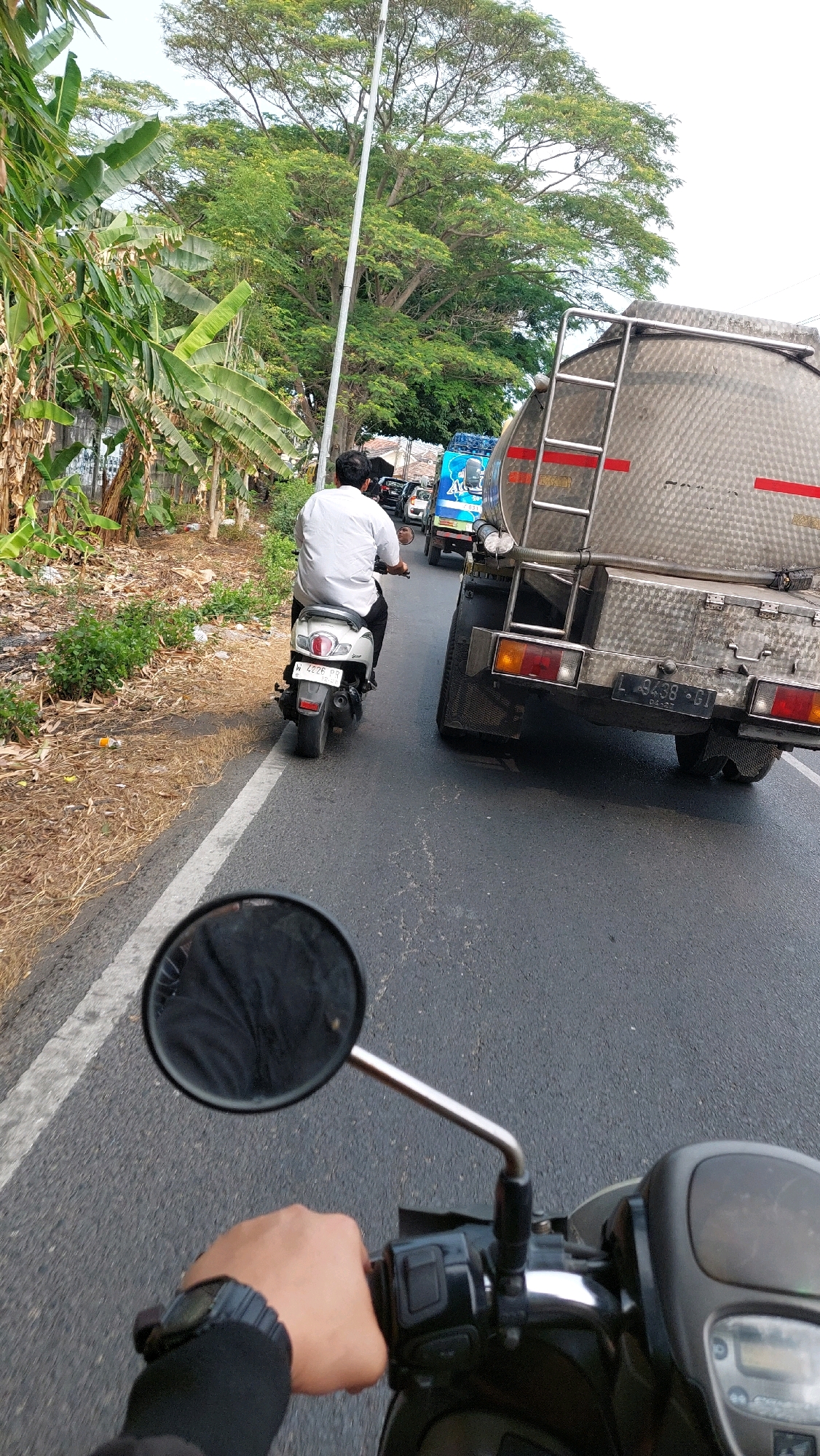The image depicts a street scene, likely in Sidoarjo, Indonesia, with multiple vehicles on a paved road. In the foreground, a person's hand is visible, gripping the handlebar of a motorcycle or scooter. A rearview mirror is attached to the handlebar, reflecting a dark surface, possibly clothing. The rider is wearing a watch on their left wrist.
Further down the road, a white scooter is being ridden by a person wearing a white shirt. This scooter has a license plate that reads "W 4226 QR".
To the right of the scooter is a large, silver tanker truck. Its rear is visible, with a license plate reading "L 9438 GT". The truck has a ladder attached to its side and appears to be a commercial vehicle.
In the background, other vehicles, including a blue tanker truck, are visible in traffic. The surroundings include lush green trees, a tall utility pole with wires, and some vegetation along the side of the road, including banana trees and dry grasses. The overall environment suggests an outdoor, daytime setting on a road with moderate traffic.