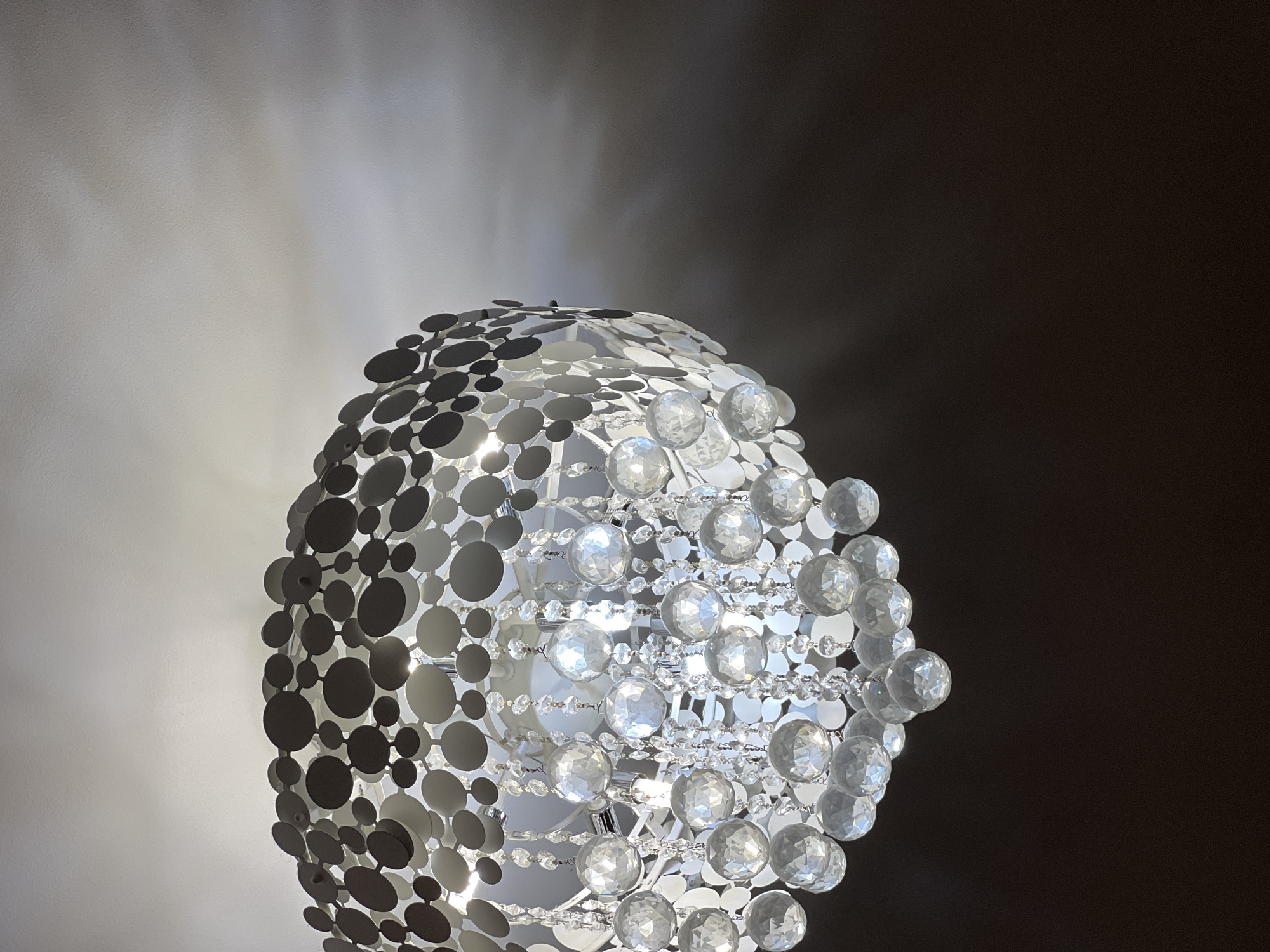 A close-up, low-angle shot focuses on a modern, circular chandelier with a multi-layered design. The outer layer consists of numerous overlapping, dark gray, circular discs attached to a white frame. Cascading down from this frame are strings of clear, faceted crystal spheres of various sizes, interspersed with smaller clear beads. The chandelier is illuminated from within, casting soft shadows on the white ceiling above and creating a gradient of light and shadow on the dark wall behind it. The location of this scene is unknown, Russia.