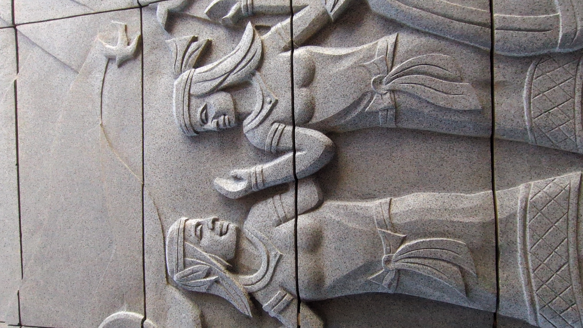 Close-up of a stone carving composed of multiple blocks. Two figures are visible in bas-relief, possibly deities or mythological figures, depicted in profile. One figure is positioned above the other with their forms slightly overlapping. Both figures wear headdresses and ornate clothing. A small bird or flying object is visible in the upper left corner of the scene. The carving is installed on an exterior wall in the city of Thành phố Tuy Hòa, Vietnam. The overall color of the scene is grey.
