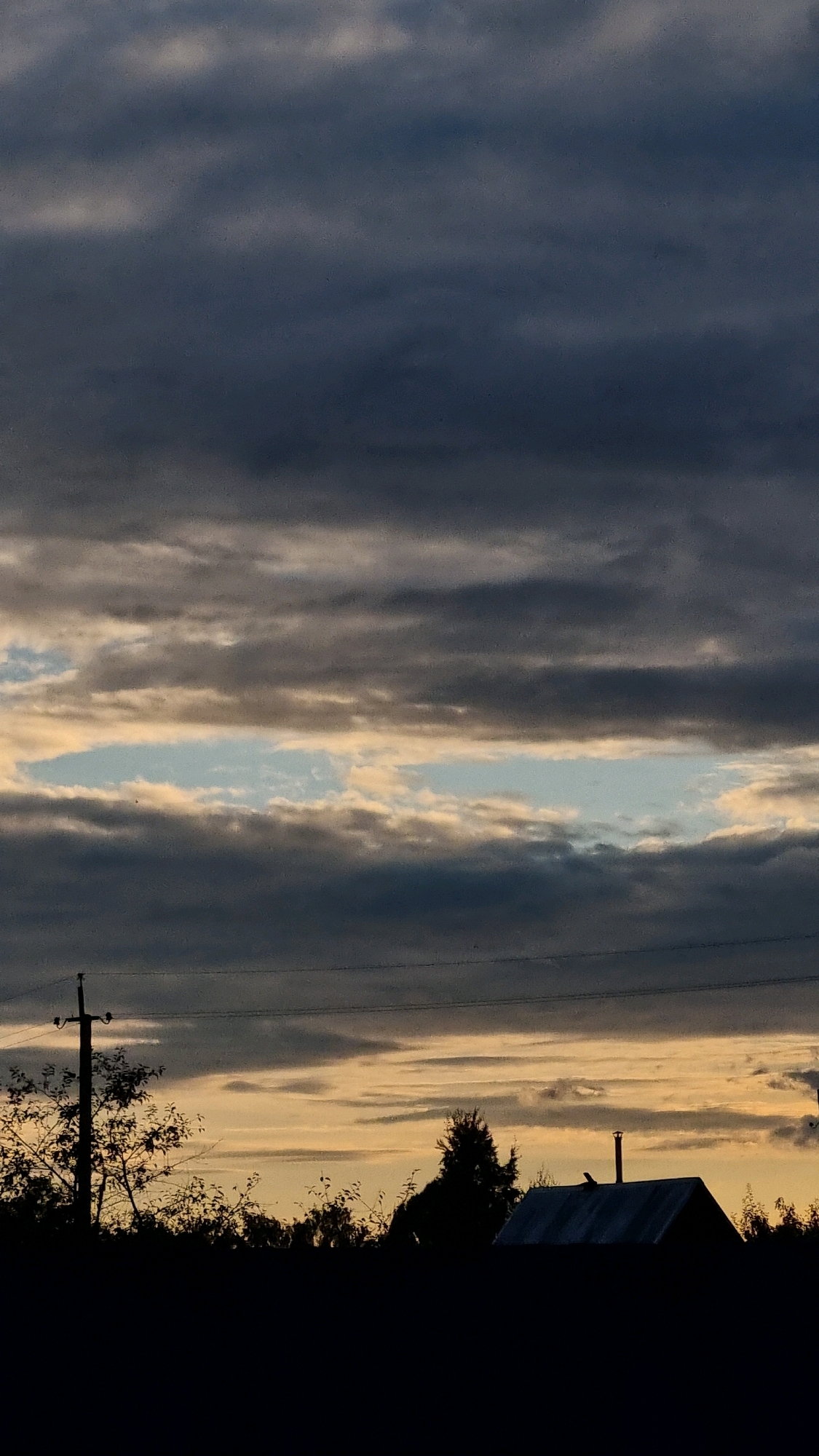 The image depicts a silhouetted rural landscape against a dramatic, cloudy sky. In the foreground, the dark outlines of trees, bushes, and a house with a prominent chimney are visible. A utility pole with wires stands to the left, further emphasizing the rural setting. The sky is filled with layers of clouds, with a horizontal band of lighter sky, showing hints of blue and yellow, piercing through the darker, heavier clouds. The overall impression is that of a moody, late afternoon or early evening in a quiet, possibly agricultural, area. The location is identified as Vysokoe, Russia.