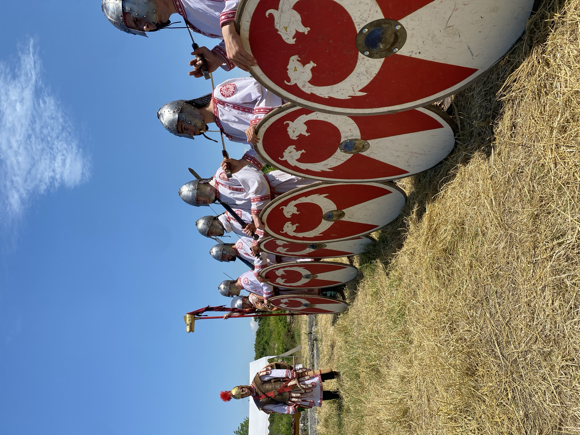 An outdoor scene in Gigen, Bulgaria, displays a historical reenactment under a clear blue sky with sparse wispy clouds. In the foreground and midground, a group of seven individuals in historical attire is arranged on a field of dry, light-brown grass mixed with green patches.
On the left, a single figure stands facing right, dressed in a golden helmet with a prominent red plume, segmented body armor over a white tunic with red trim, and red leg coverings. This individual holds a gladius-style sword in their right hand, pointed downwards.
To the right, a line of six figures stands facing left, positioned in a staggered formation. Each wears a silver, conical helmet and a white tunic with red embroidered patterns around the collar and sleeves. The figures hold what appear to be spears or long swords. The second figure from the left in this line holds a standard featuring a golden animal head, possibly a dragon (Draco), atop a red pole with a trailing red banner.
Five round shields, predominantly red with a large white central design resembling two stylized opposing dragons or serpents, are propped upright in the immediate foreground, held by the line of figures. Each shield has a central metal boss.
In the background, behind the single figure, a white tent is visible on the left, partially obscured by distant green foliage and a darker strip of ground. The scene is brightly lit by direct sunlight.