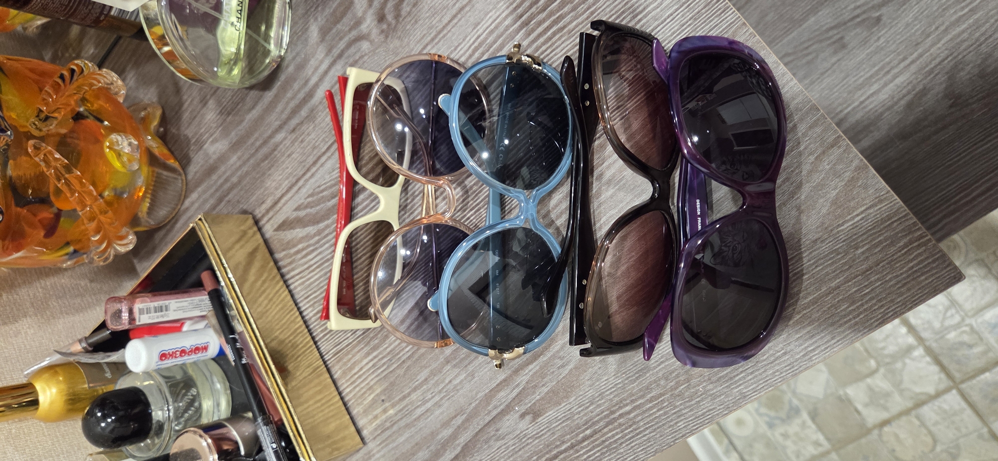The image depicts a collection of sunglasses and cosmetics laid out on a wooden surface, possibly a table or dresser. From top to bottom, the sunglasses are a pair with a cream and red frame, followed by a pair with light brown round lenses and frames, then a pair with light blue round lenses and frames, a pair with brown gradient lenses and black frames, and finally a pair with dark purple frames and lenses. In the upper left corner, a gold-trimmed tray holds various cosmetic items, including a clear bottle with a black cap, a gold-capped jar, and several lip liners or pencils. To the right of the tray, an ornate glass object with orange and yellow hues is visible, and further to the right, a round glass bottle of perfume with a green liquid and a label that reads "CHANEL" is present. The overall setting suggests a personal vanity or dressing area. The specific location of Lipetsk, Russia, is not discernible from the visual information provided in the image.