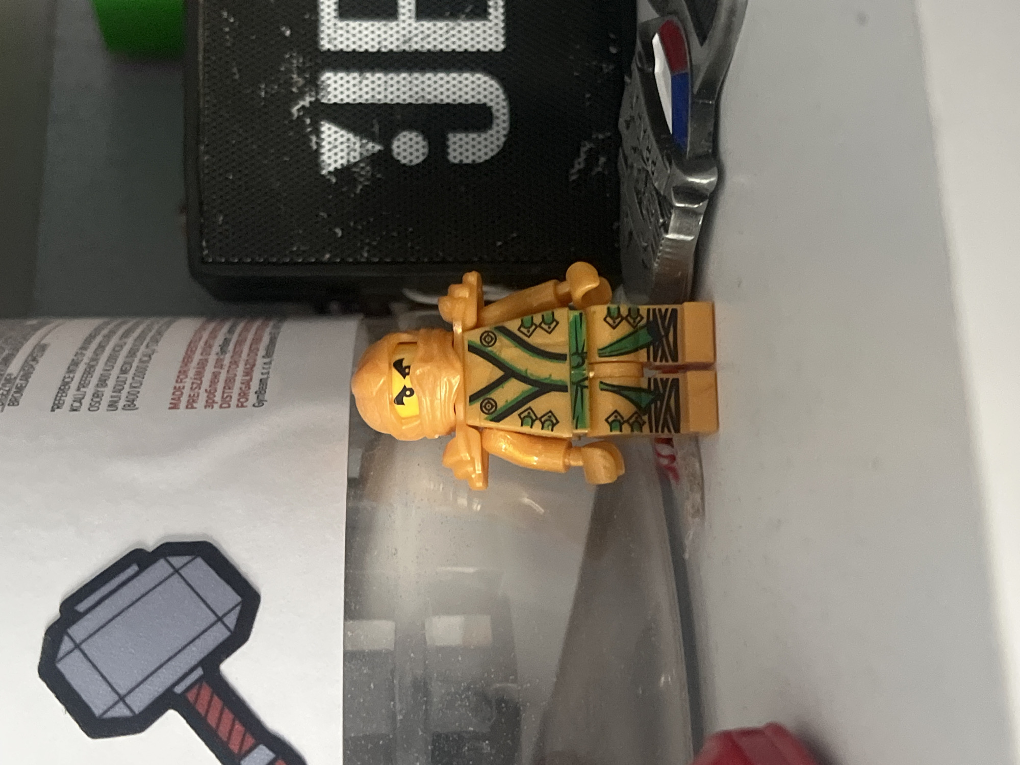 A Lego minifigure of the ninja Cole, in his golden attire from the Ninjago Masters of Spinjitzu television series, stands on a white shelf. The minifigure is positioned in the center of the frame, with its left side facing the viewer. Behind the minifigure, a black JBL speaker is visible, with the white JBL logo prominently displayed. To the left of the speaker, a cylindrical white bottle with text is partially visible. Further to the left, a stylized graphic of Thor's hammer, Mjolnir, is imprinted on the bottle. In the foreground, a portion of a silver medal with red, white, and blue stripes is present. The background is a light gray wall, suggesting an indoor setting. There are no people or explicit actions depicted in the scene. The setting is not identifiable as being in Pokrovnik, Bulgaria.
