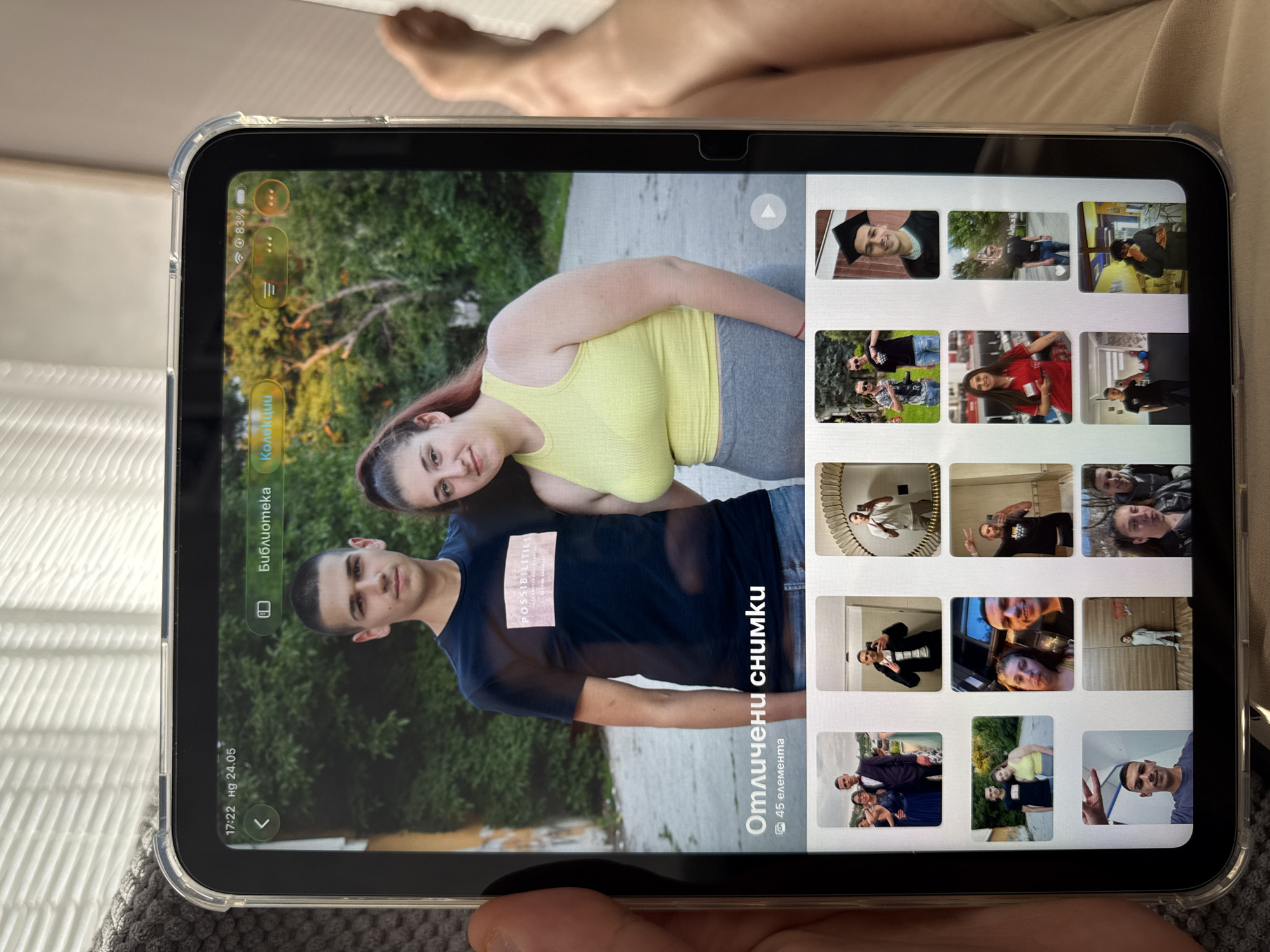 A medium shot depicts a tablet displaying a photo album interface. The main image featured is a couple posing for a photo outdoors amidst greenery, possibly in a park. The man wears a dark blue t-shirt, while the woman sports a yellow tank top and grey pants. To the side of the main photo, a grid of smaller images hints at other photos in the album, featuring both individuals in various settings, including graduation and social events. The interface is in a language other than English, indicating a regional adaptation. The tablet is held by a person, suggesting the viewer is looking at the screen through the perspective of the person holding the tablet. Based on the text displayed, the photo album is called "Отличени снимки" (Featured photos), and it has 45 photos. The date on the screen reads "17:22 пд 24.05". The location of Lovech, Bulgaria is not inferable from the visual elements of the image.