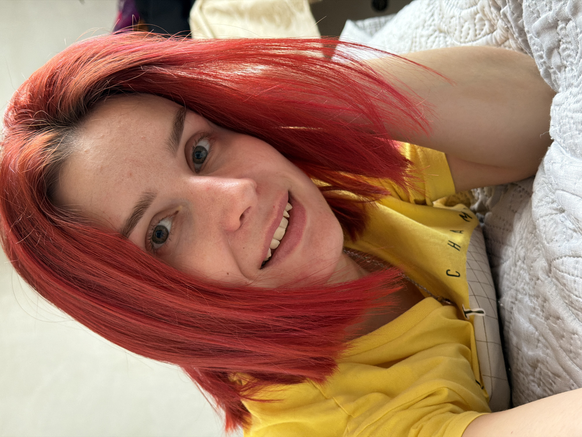 A fair-skinned woman with red hair is lying down, smiling at the camera. She is wearing a yellow t-shirt. She appears to be lying on a bed with white bedding. The location is not discernible from the image; there is no indication it is in Chaikovsky, Russia.