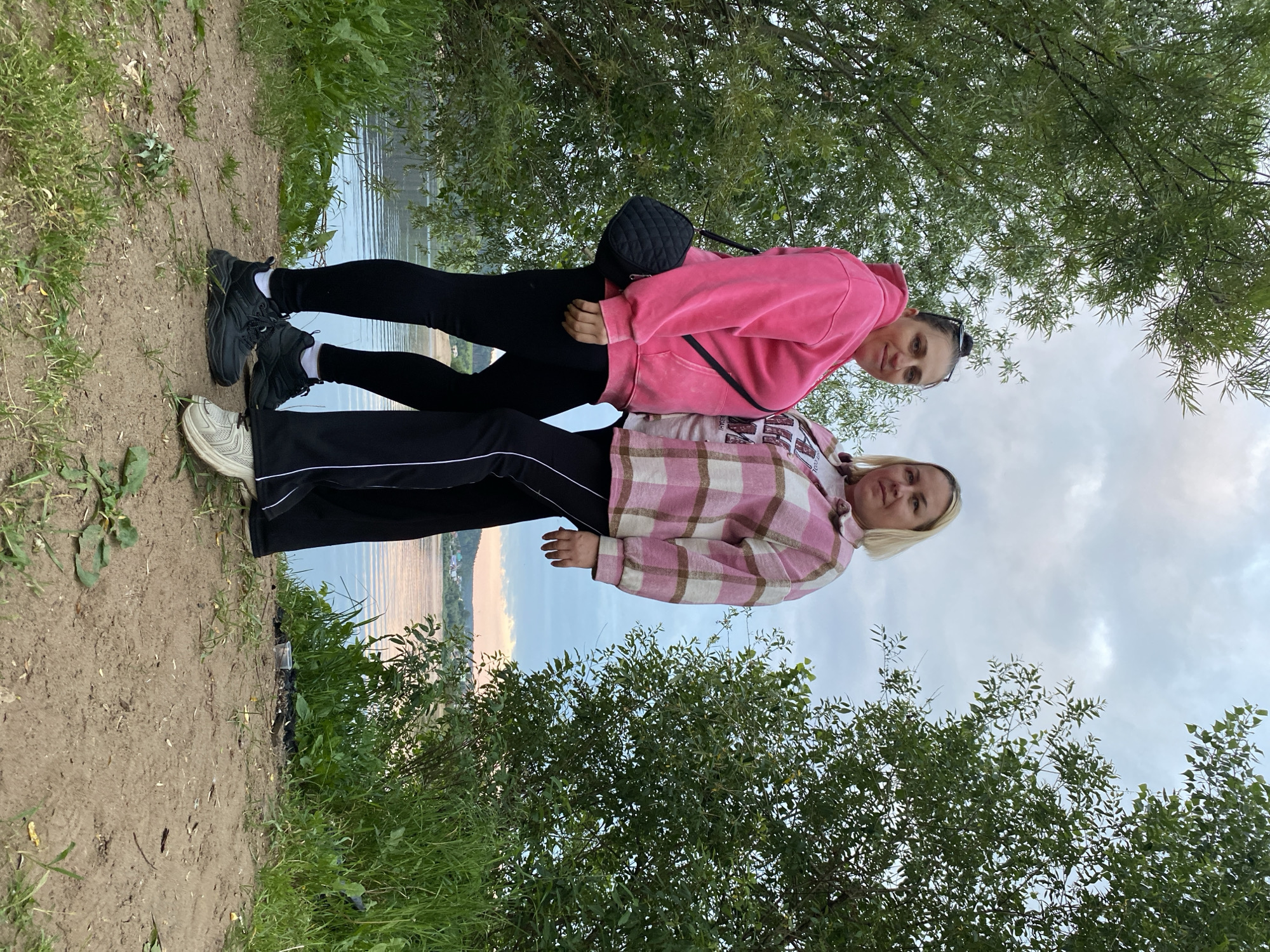 Two women are standing on a dirt path next to a body of water, with trees on either side and a cloudy sky above. The woman on the left is wearing a pink hooded sweatshirt and black leggings, and a black quilted purse is slung across her body. The woman on the right is wearing a pink and white plaid shirt jacket over a t-shirt and black pants with a white stripe. They are both wearing athletic shoes. The background shows a body of water, with a distant shoreline and some buildings visible. The location is on the shore of the Volga River near the village of Noviy, Nizhny Novgorod Oblast, Russia.