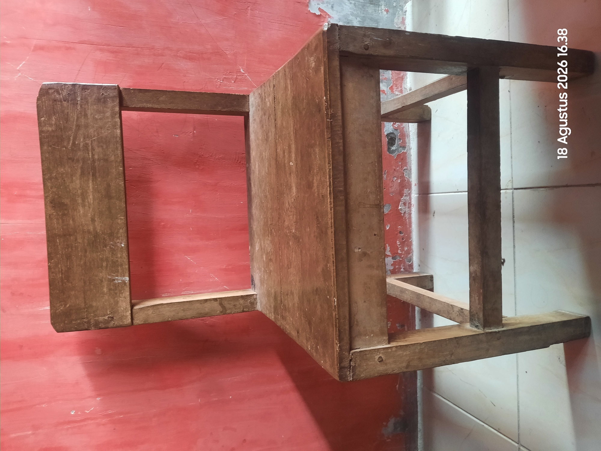 A medium shot captures a rustic wooden chair situated in front of a textured, salmon-pink wall. The chair, made of aged wood, exhibits signs of wear, including scratches and a faded finish. It has a simple design with four legs, a seat, and a backrest. The wall behind the chair is uneven and displays patches of peeling paint, revealing underlying layers. The floor beneath the chair is tiled with a light-colored, square pattern. The scene is illuminated by natural light, casting a shadow of the chair onto the floor. In the bottom right corner, a timestamp reads "18 Agustus 2026 16.38".