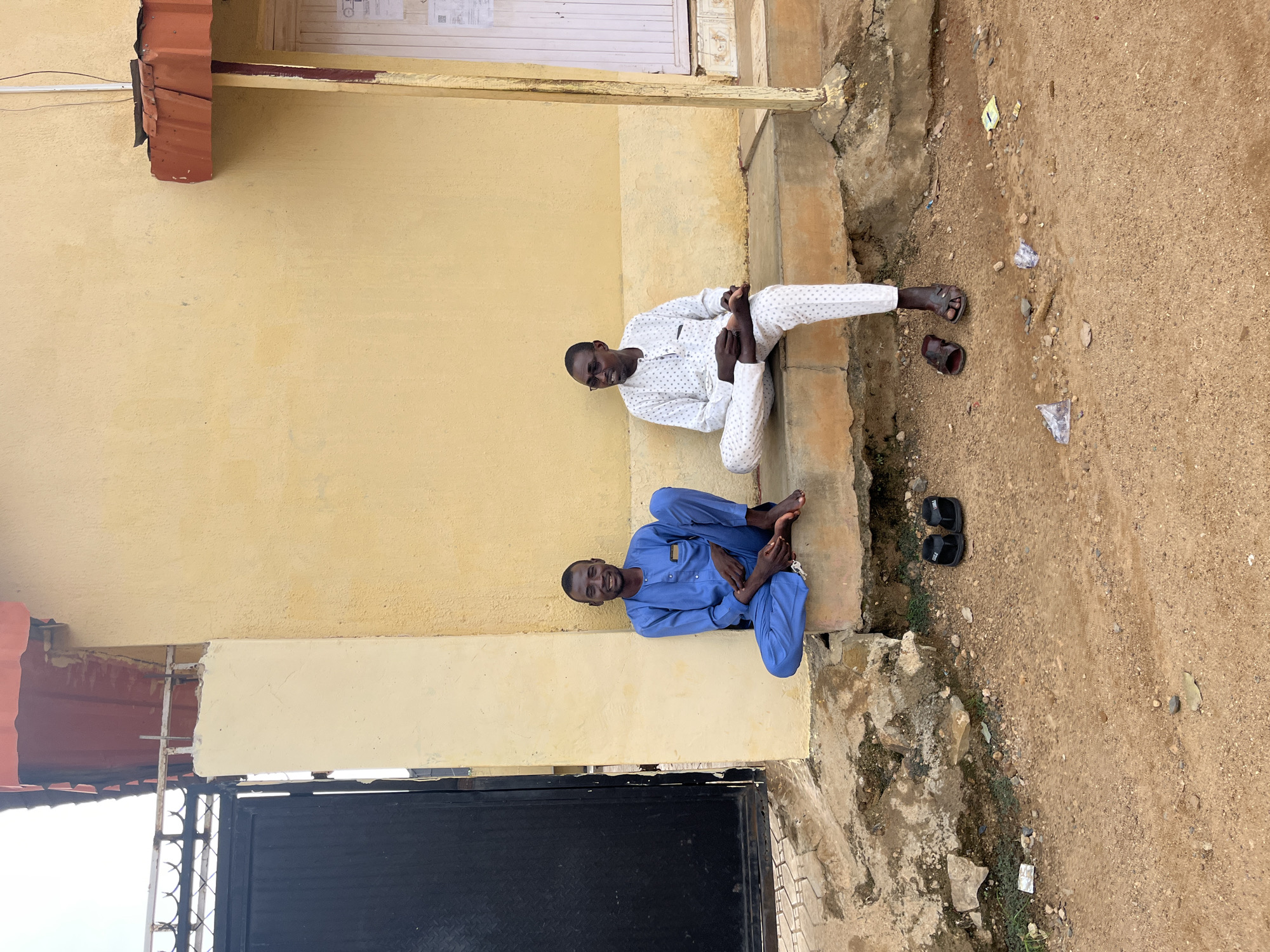 Two men are sitting on a concrete ledge outside a yellow building in Jos, Nigeria. The man on the left is wearing a blue outfit and the man on the right is wearing a white outfit with a pattern. Both men are barefoot. In front of them on the ground are two pairs of sandals. Behind them is a black metal gate. The building appears to be a simple, one-story structure with a corrugated metal roof. The ground is covered in dirt and gravel, with some scattered debris. The overall scene suggests a casual, outdoor setting in an urban environment.
