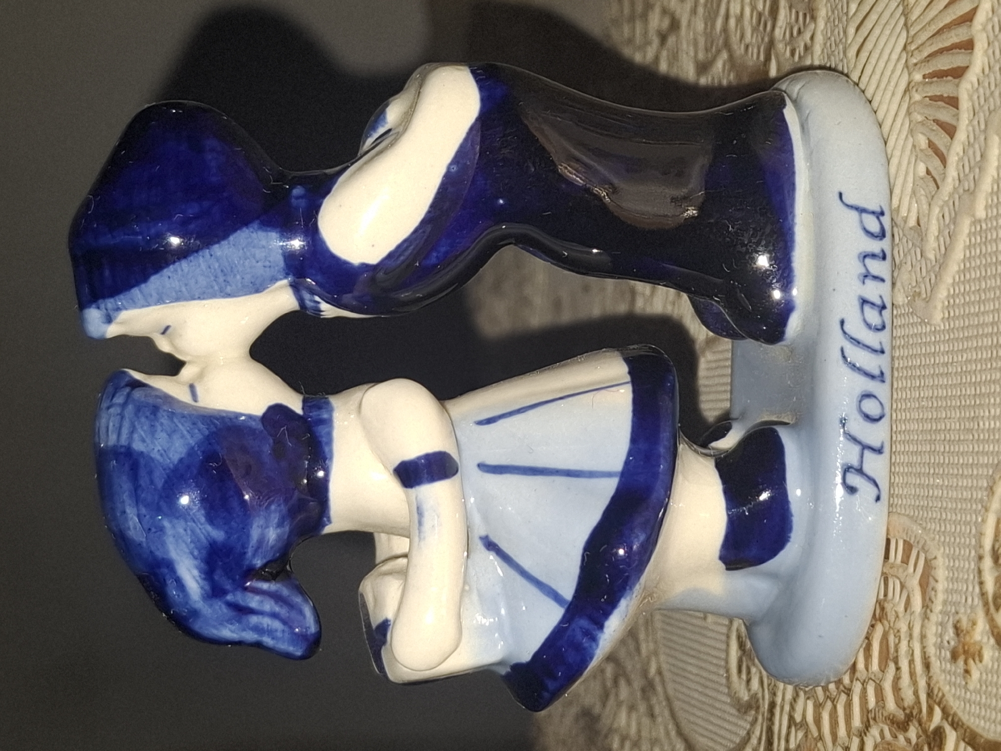 The image shows a ceramic figurine of two children in blue and white attire, leaning in for a kiss. The figurine is placed on a light blue base, which has the word "Holland" inscribed in blue cursive. The figurine is positioned on a textured surface with a patterned fabric underneath. The background is dark and appears to be a wall. There is no indication of the city of Almaty, Kazakhstan, in the image.