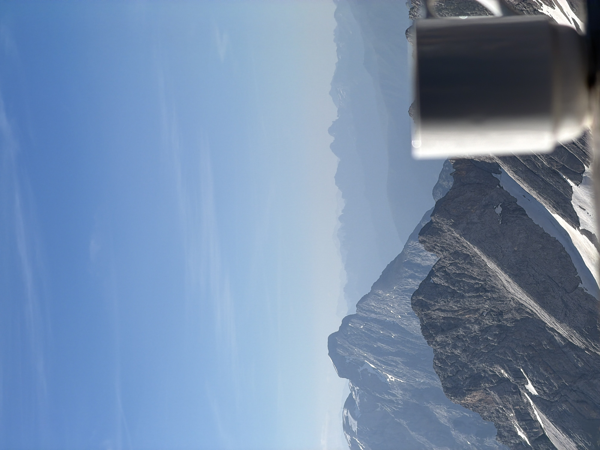 The image shows a white mug in the foreground, out of focus, with a panoramic view of the Alps in the background. The mountains are snow-capped and rugged, with a clear blue sky above. The scene suggests a serene moment, possibly a break during a hike or a quiet morning with a view. The location is likely the Zugspitze, Germany's highest peak, which is accessible from Garmisch-Partenkirchen.