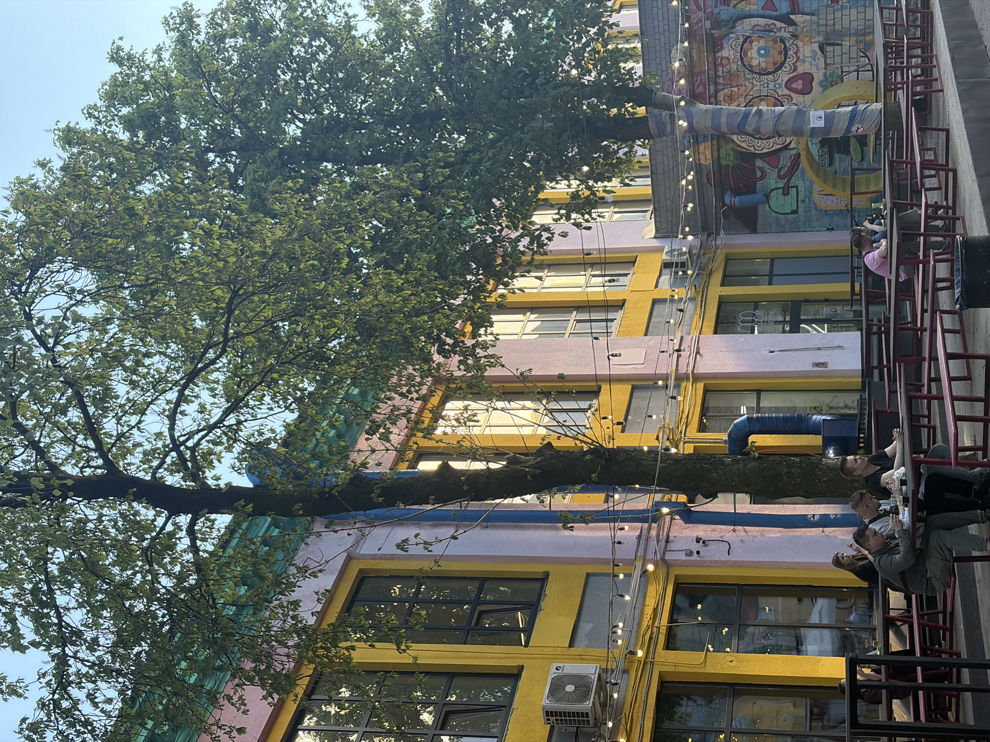 The image shows an exterior view of a colorful, multi-story building, likely in Minsk, Belarus. The building has alternating levels of yellow and lavender paint. It features windows with dark frames, and an air conditioning unit is mounted on one of the exterior walls. Strings of lights are draped along the building's facade. A mature tree with dense green foliage is positioned prominently, its trunk extending horizontally in front of the building. Adjacent to the building is a red metal fire escape with several people sitting and standing on it. They appear to be interacting and enjoying the view. Part of the building has a mural with intricate designs.